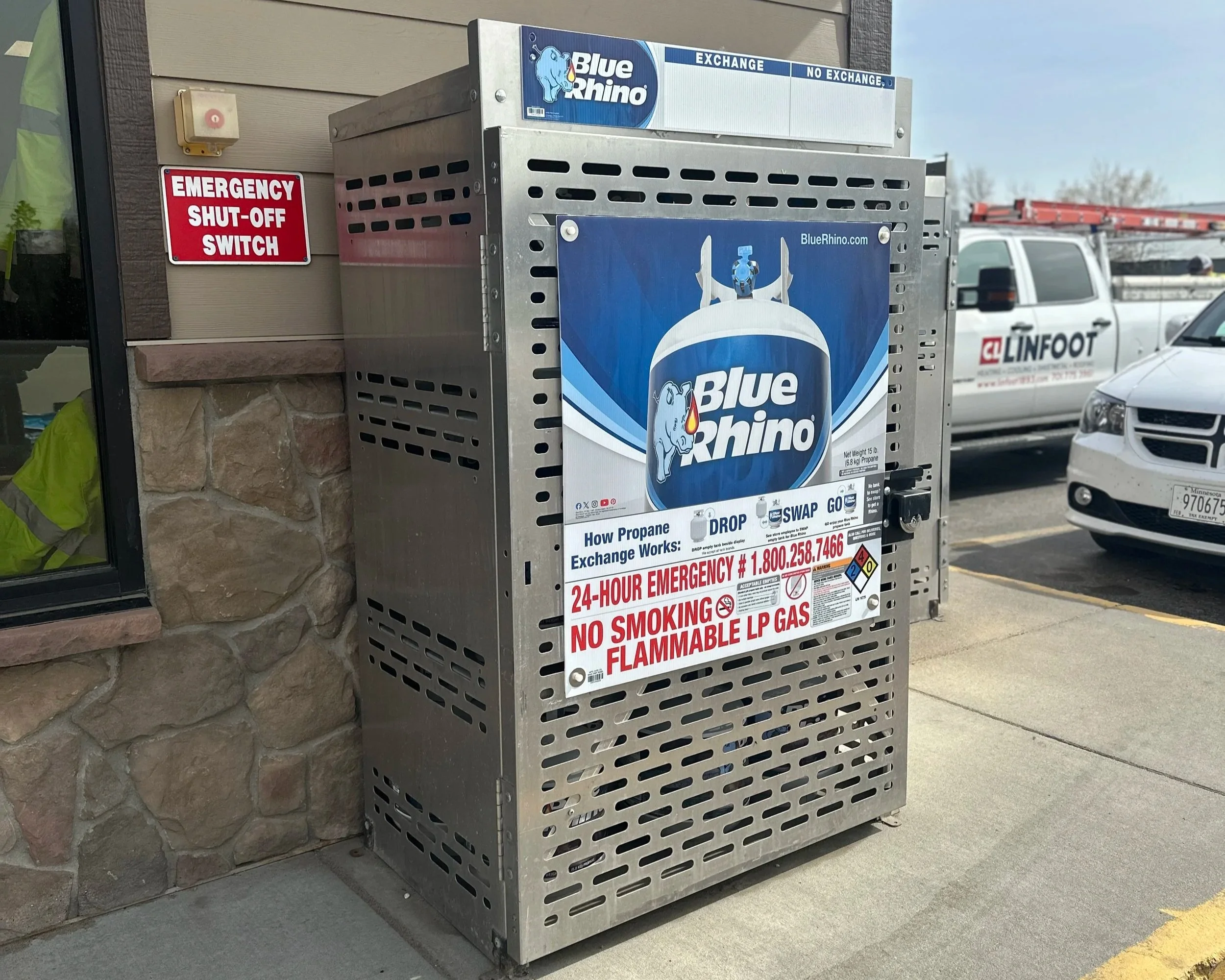 A propane exchange station outside a building. The station is made of metal with vented sides and displays a large blue sign with the Blue Rhino logo, featuring a cartoon rhinoceros. There is a notice with instructions on how propane exchange works, along with emergency contact information. To the side, there is a red emergency shut-off switch on the wall, and parked vehicles are visible in the background, including a white truck and a white car.
