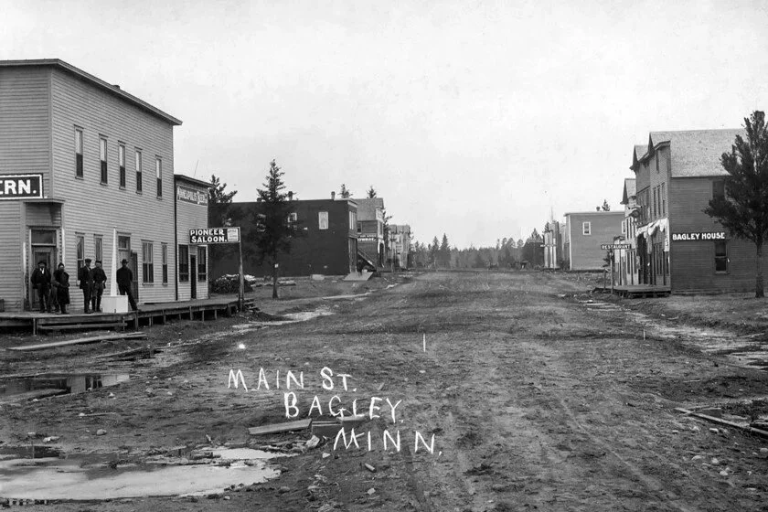Historic black-and-white photo of Main Street in Bagley, Minnesota, with dirt road, early 20th-century buildings, and a few people standing outside.