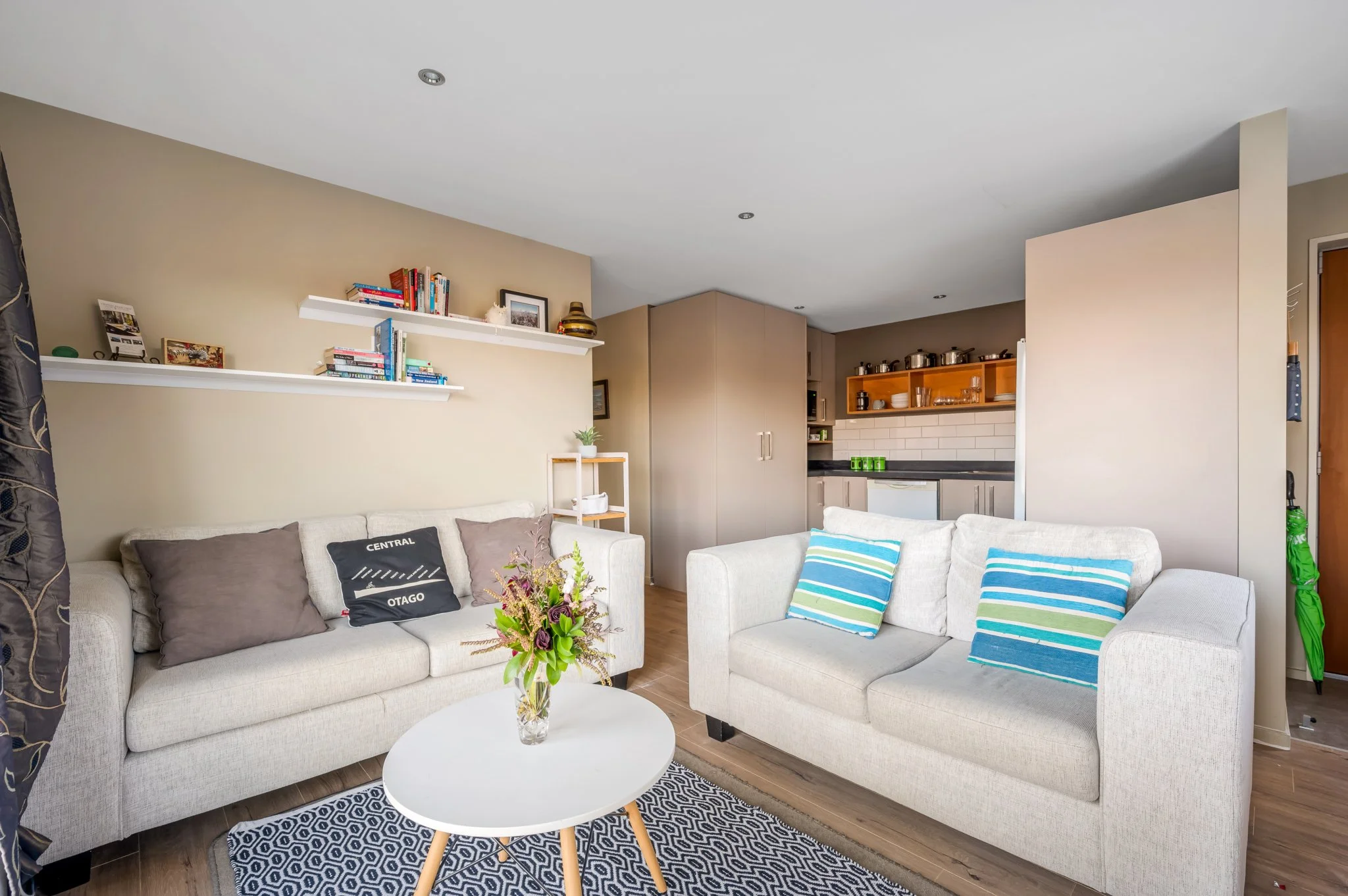 Cosy living room with two white sofas, a patterned rug, a small white coffee table with a vase of flowers, floating shelves, and an open kitchen area with wooden cabinets and black countertops.