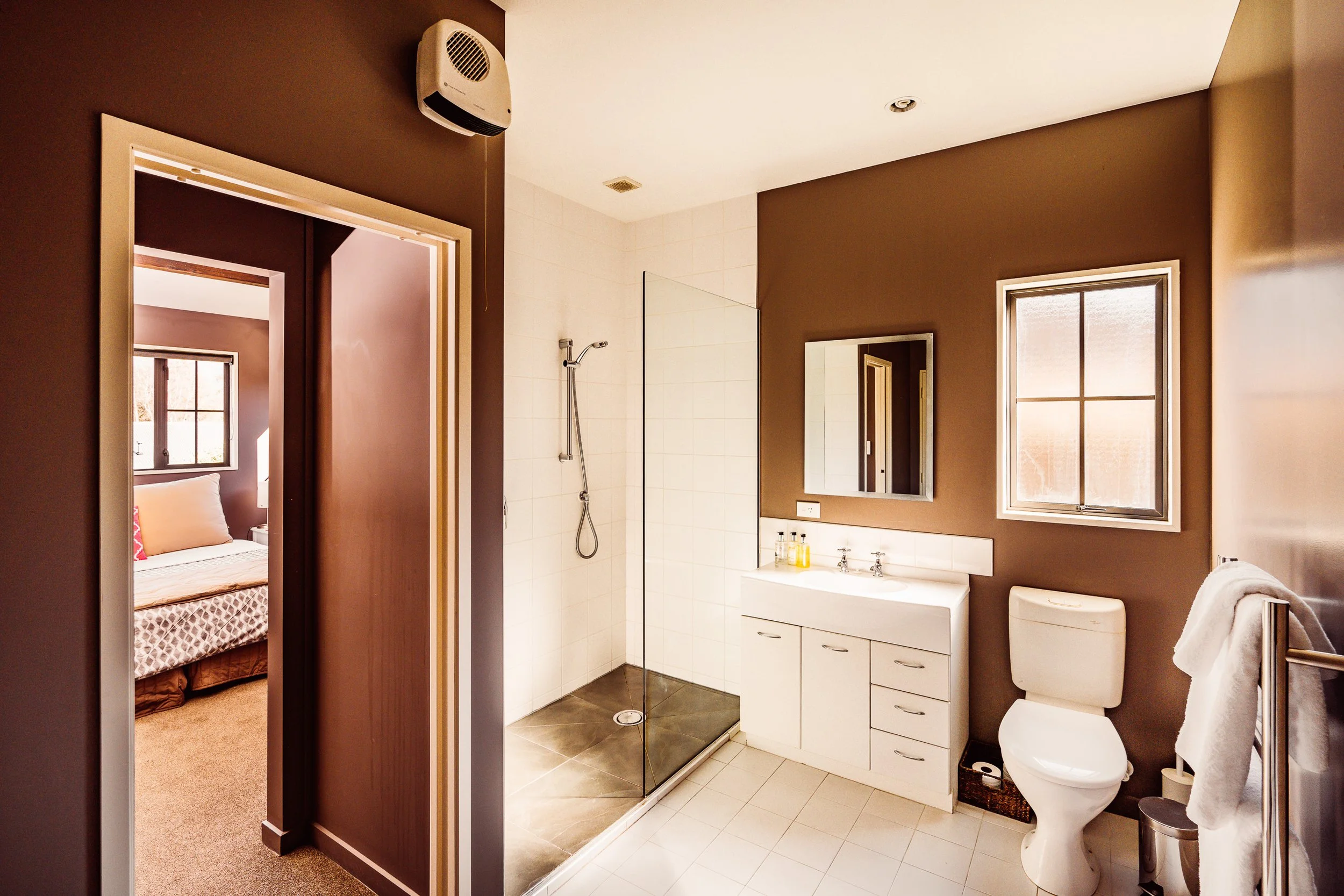 Bathroom with a shower, white vanity with a mirror, toilet, and window. Bedroom visible through doorway.