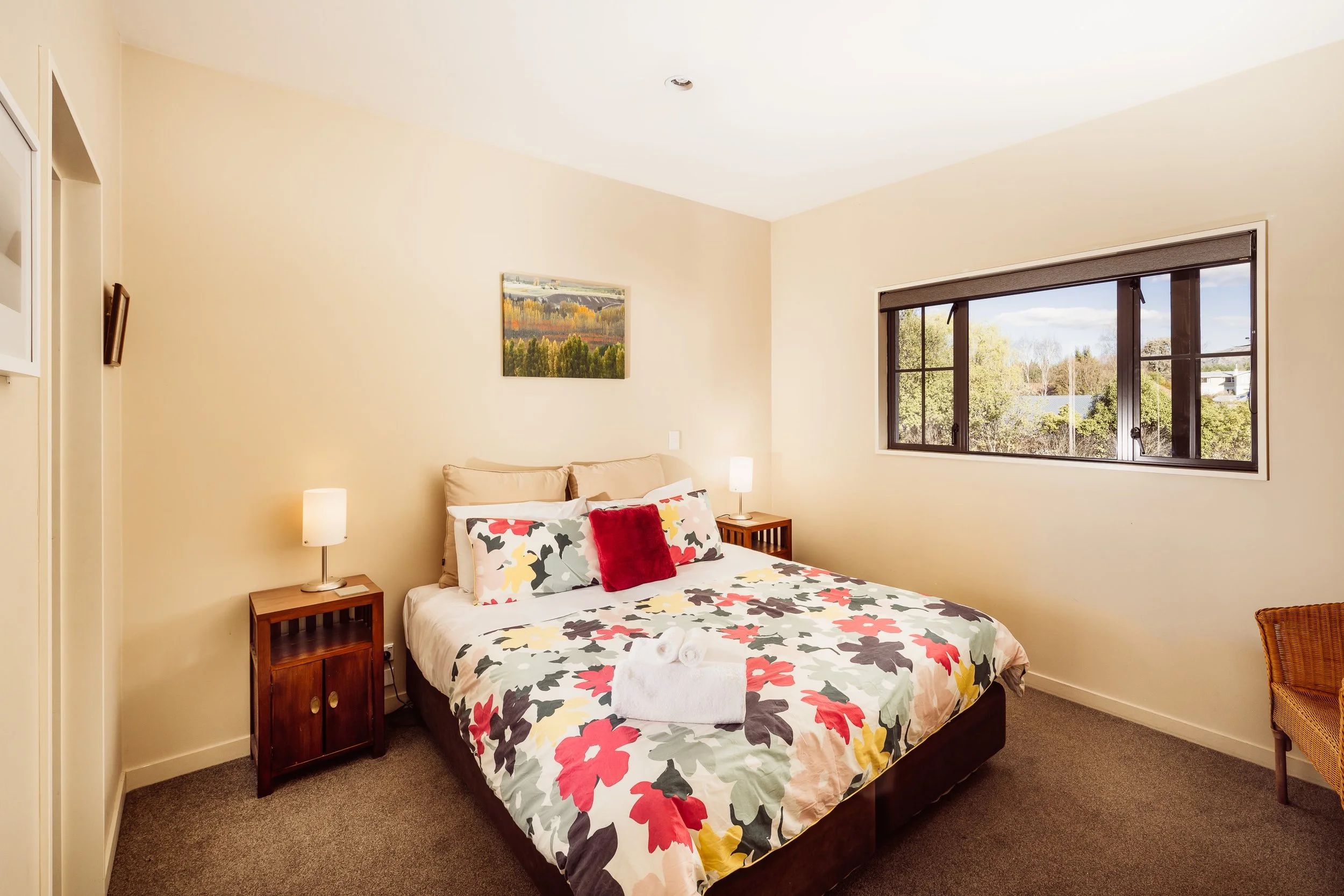 A bedroom with a bed covered in a floral duvet, two pillows, and red accent pillow, bedside tables with lamps, a window showing outdoor greenery, and a painting on the wall.