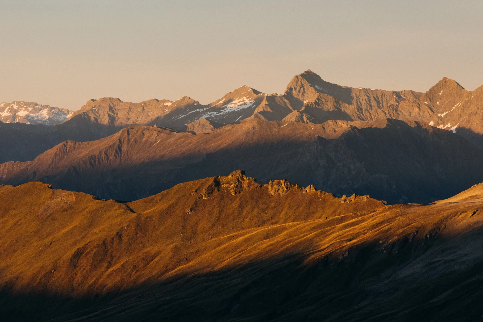 Sunset over rugged mountain landscape with snow-capped peaks in the distance and rolling grassy hills in the foreground.