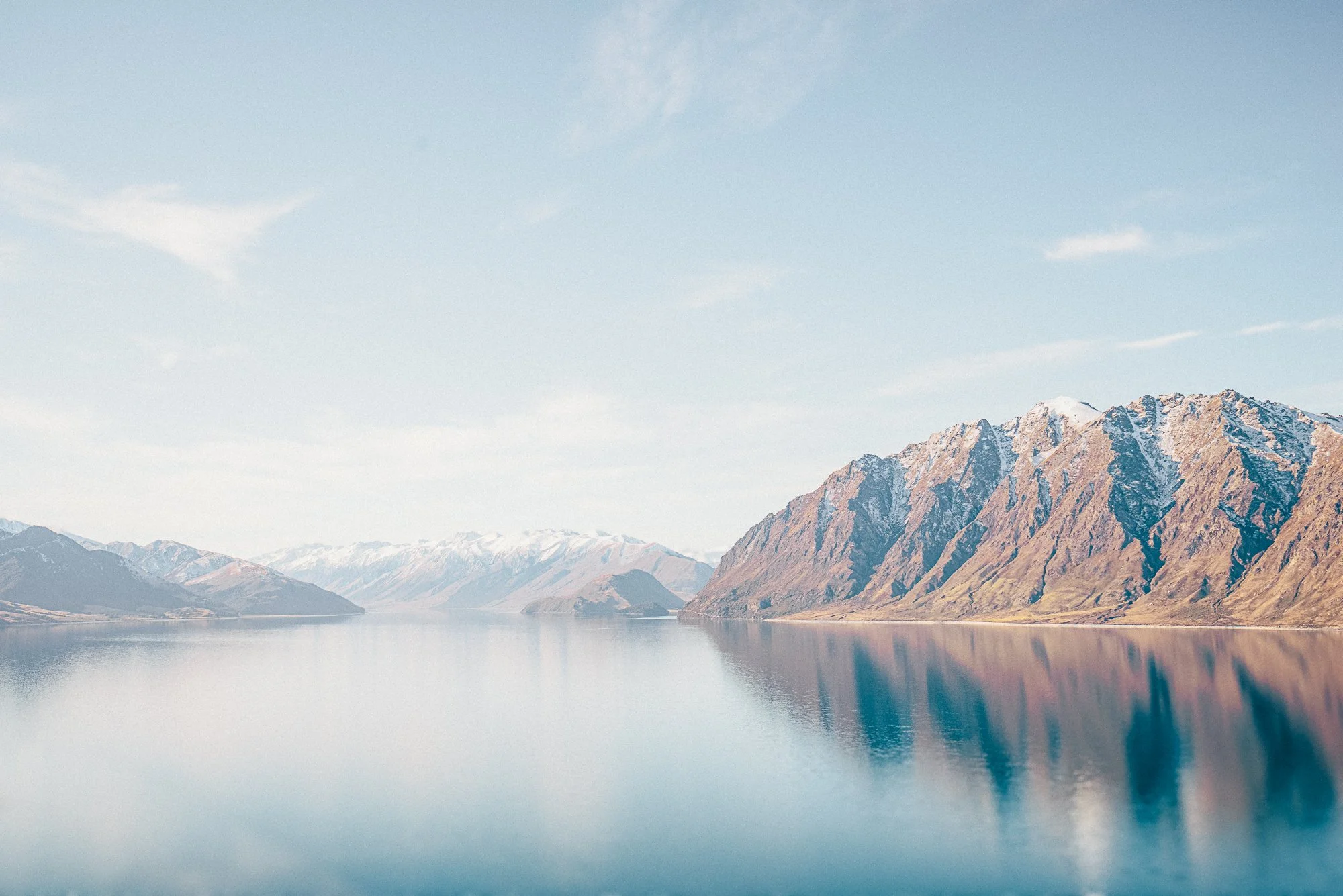 Lake Hawea reflecting mountains on either side with snow caps, under a partly cloudy sky.