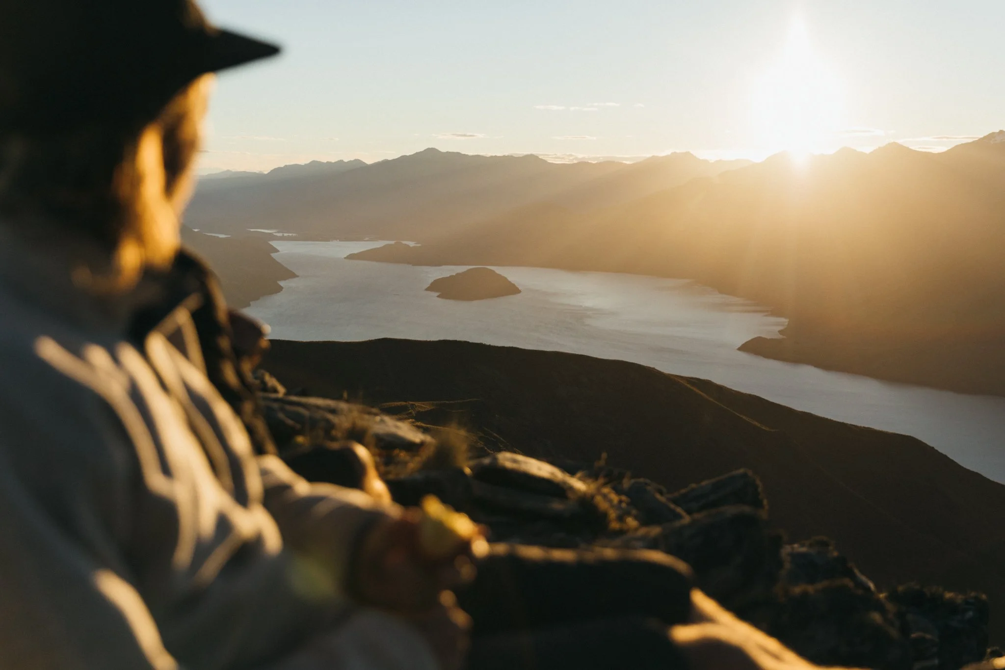Person with a hat and outdoor gear sitting on a mountain at sunset, overlooking a river and hills in the distance.