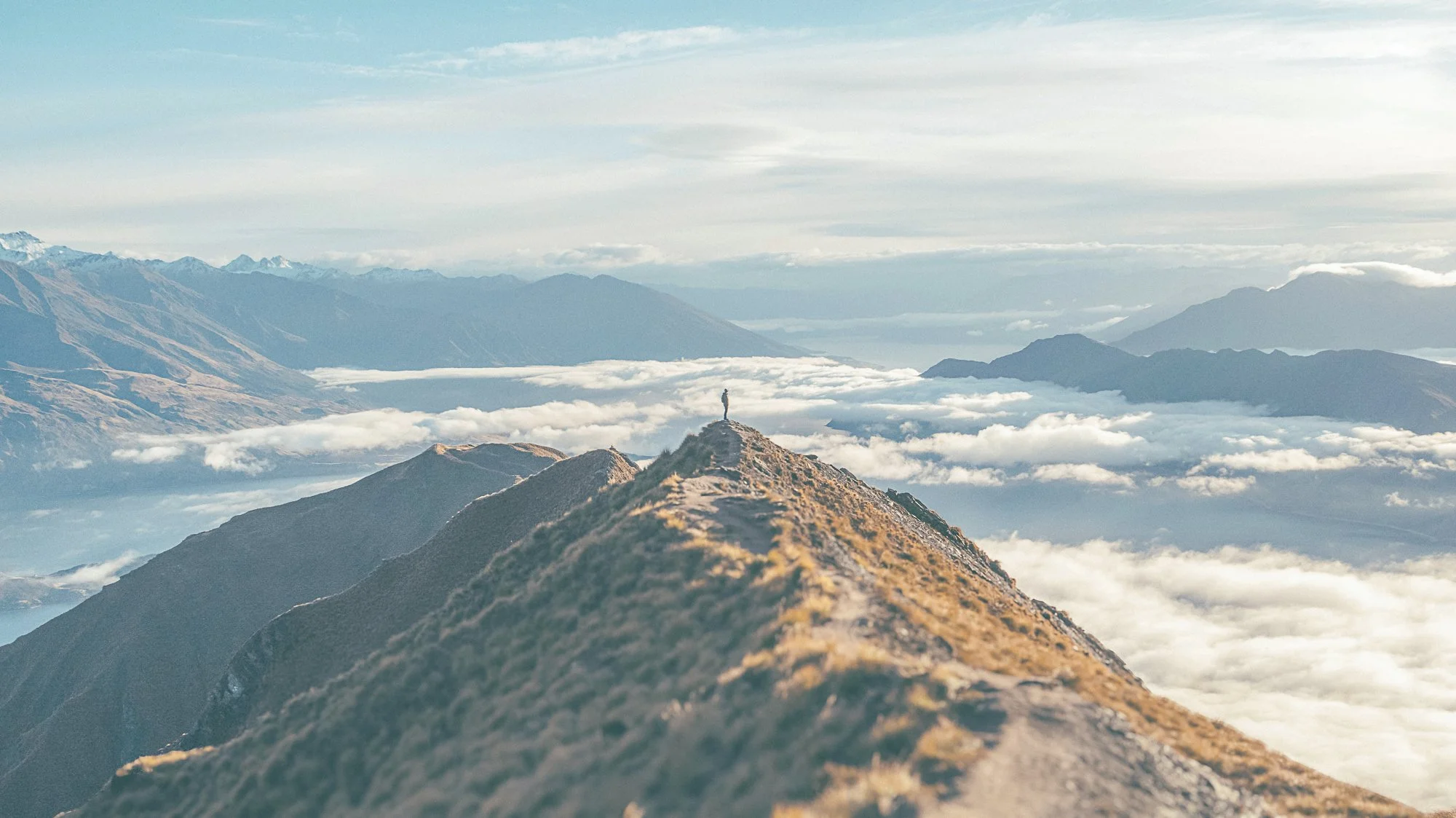 A person standing at the summit of a mountain ridge overlooking a valley filled with clouds and distant mountain ranges.