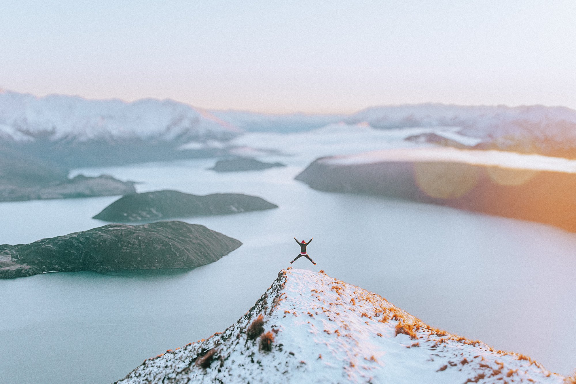 Person standing on snowy mountain peak with arms raised, overlooking a scenic lake with islands and snow-capped mountains in the background during sunrise or sunset.