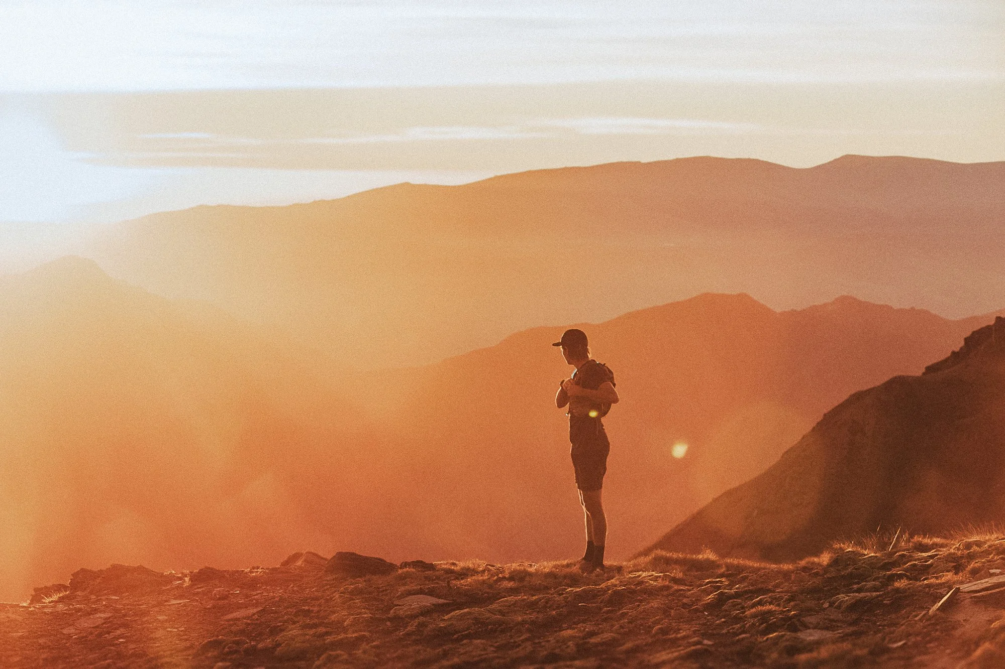 A person standing on a mountain at sunset, wearing a cap, shorts, and carrying a backpack, with a landscape of distant mountain ranges in the background.