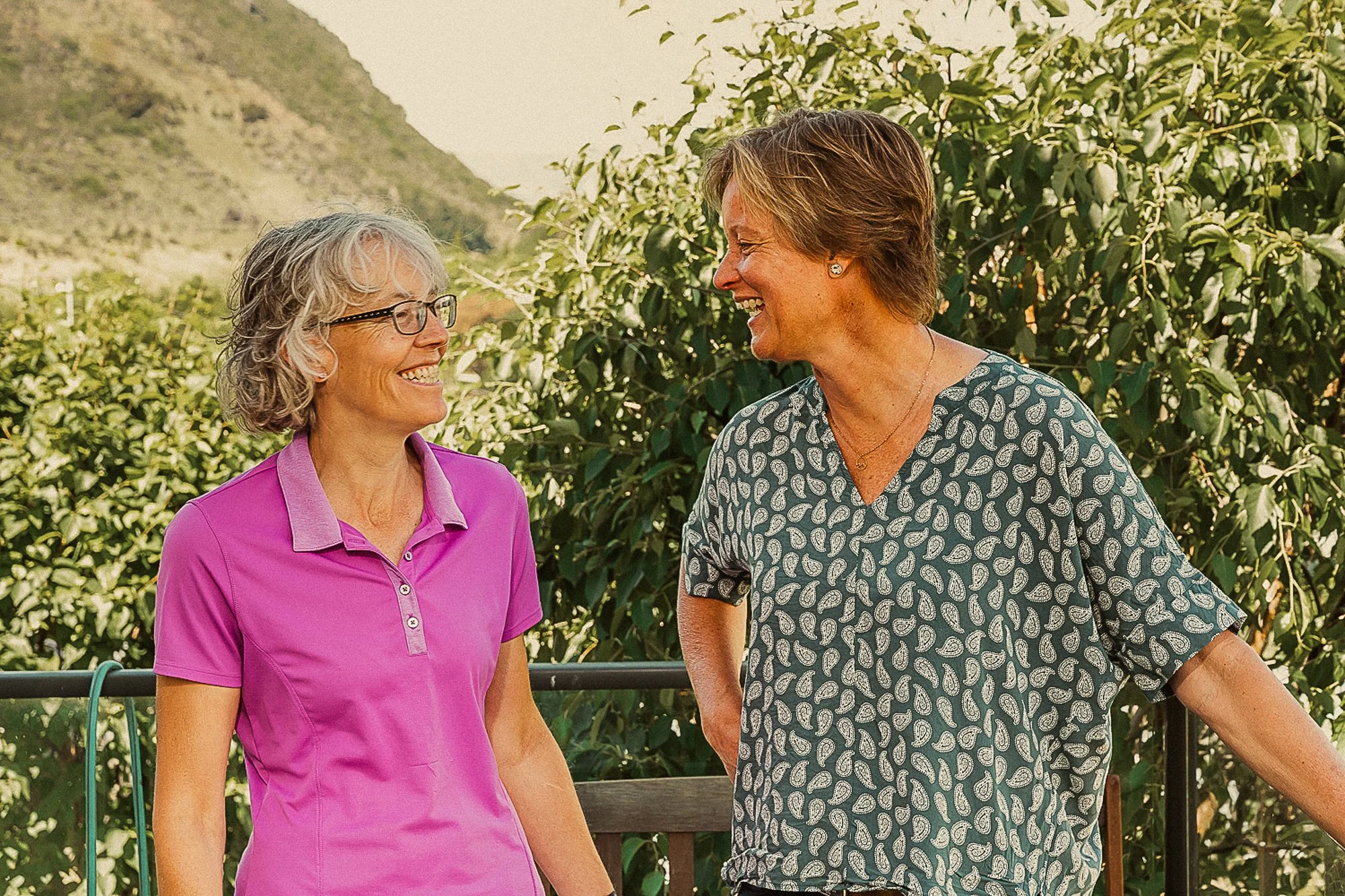 Two women smiling and talking outdoors, with lush green foliage and hills in the background.