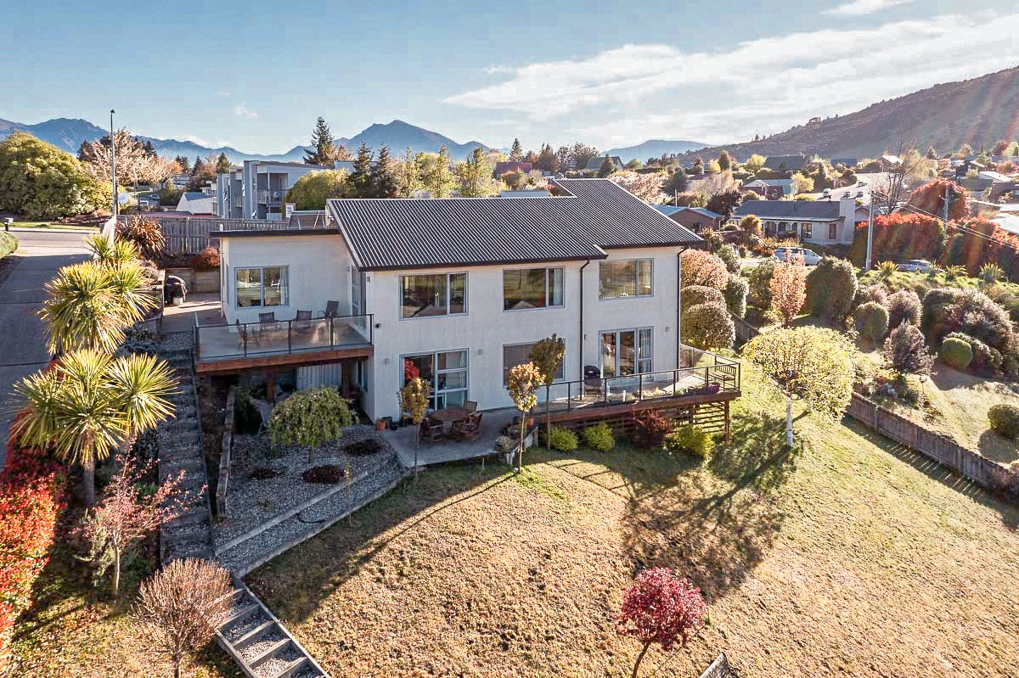 A white two-story house with a black metal roof, front and back decks, large windows, and a spacious backyard with trees, shrubs, and a lawn, located in a suburban neighborhood with mountain views.