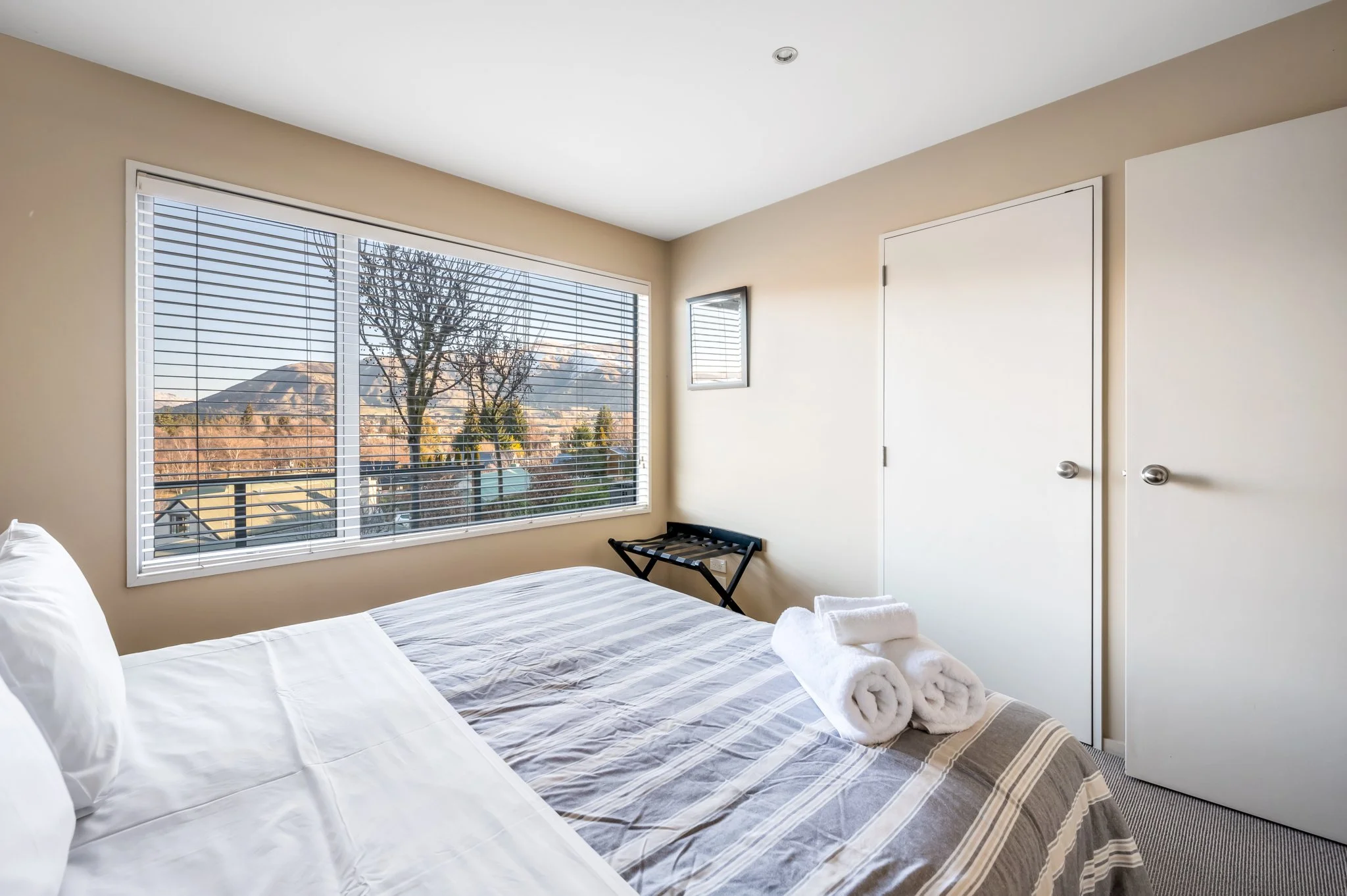 A bedroom with a large window showing mountains and trees outside. The bed has white and gray bedding with two rolled towels placed on top. There is a small black luggage rack near the window, and a small mirror is mounted on the wall. The room has beige walls and white doors.