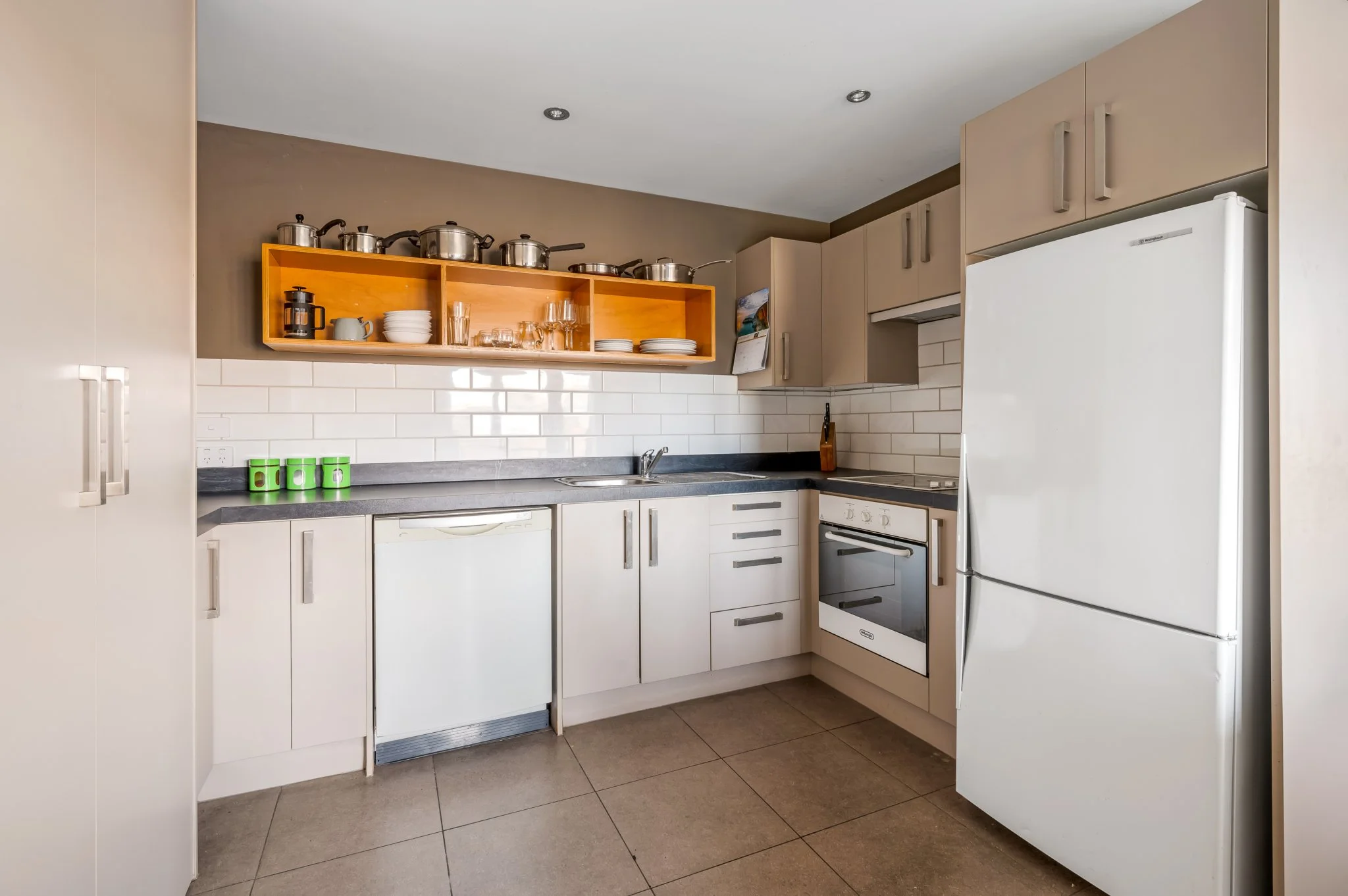 A clean kitchen with beige cabinets, a gray countertop, white backsplash tiles, and a white refrigerator. Open wooden shelf holds pots, glasses, and dishes. Three green cups are out, ready for breakfast.