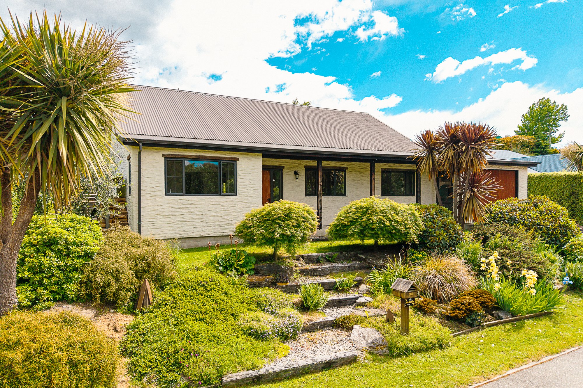 Front view of a suburban house with white brick walls, black window frames, and a gray metal roof surrounded by lush green plants and bushes.