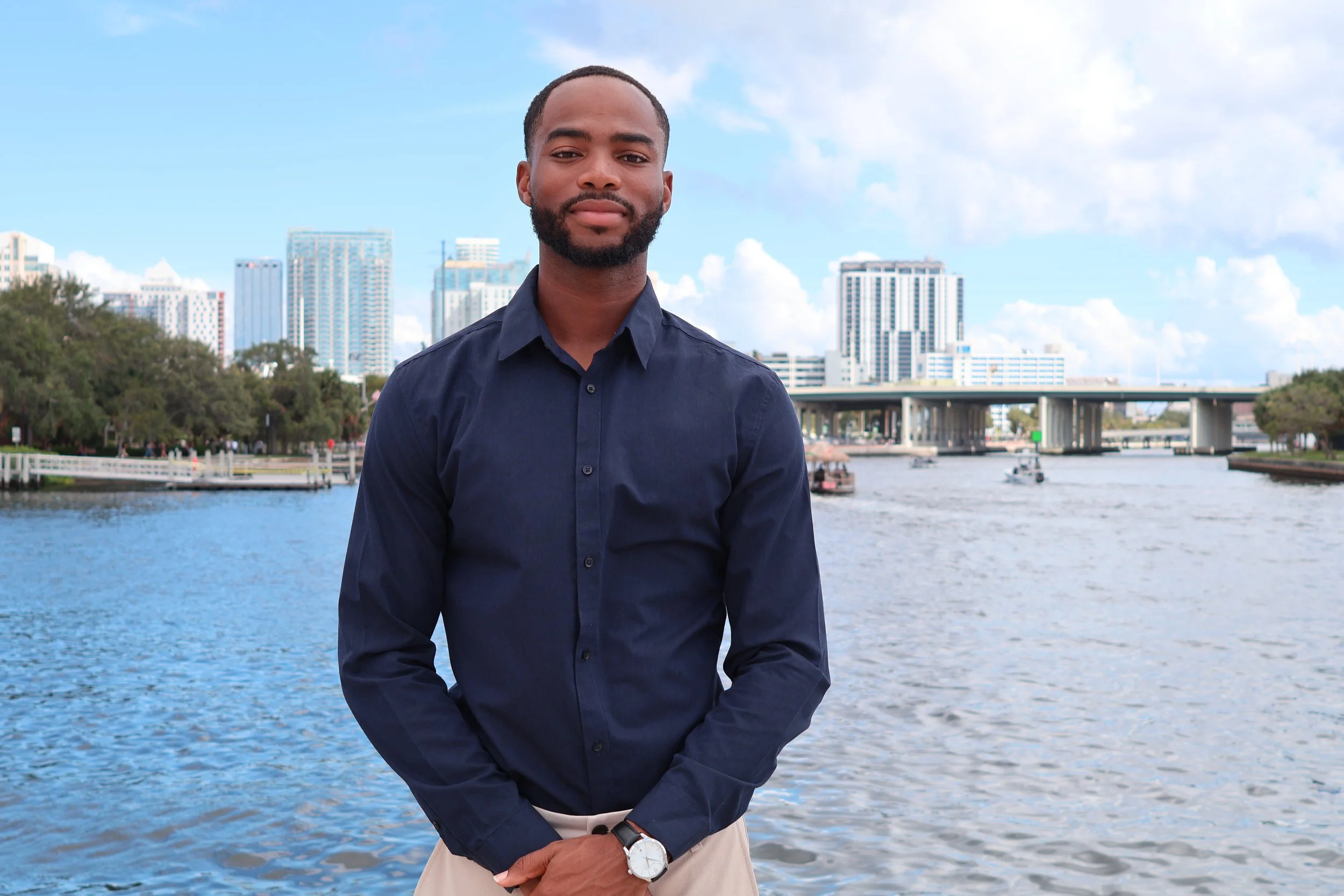 A man with a beard wearing a dark blue shirt and beige pants standing by a river with city skyscrapers in the background.
