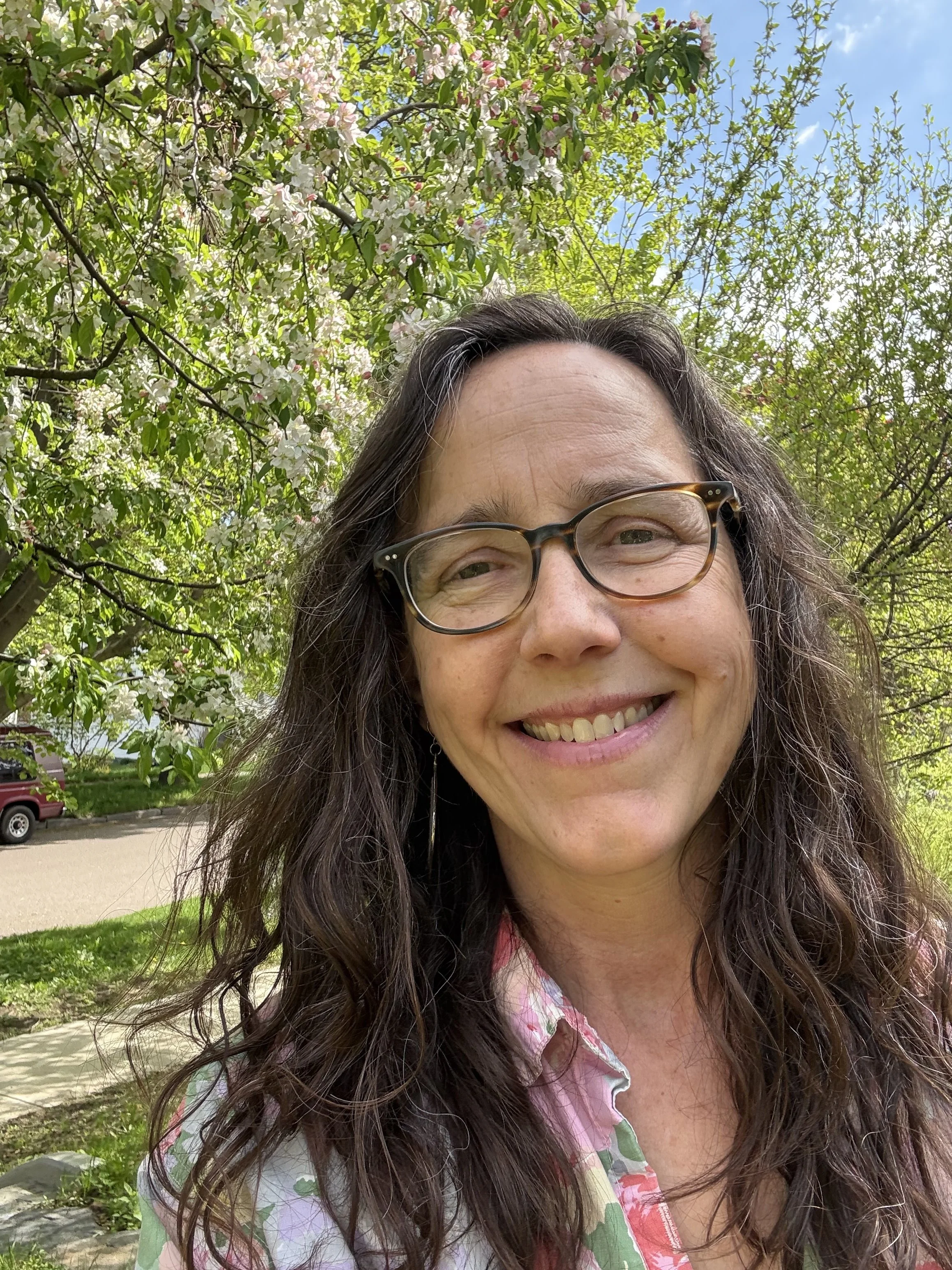 A smiling woman with long brown hair and brown, tortoiseshell glasses. She's wearing a floral-print shirt and standing outside under a crabapple tree in full bloom.