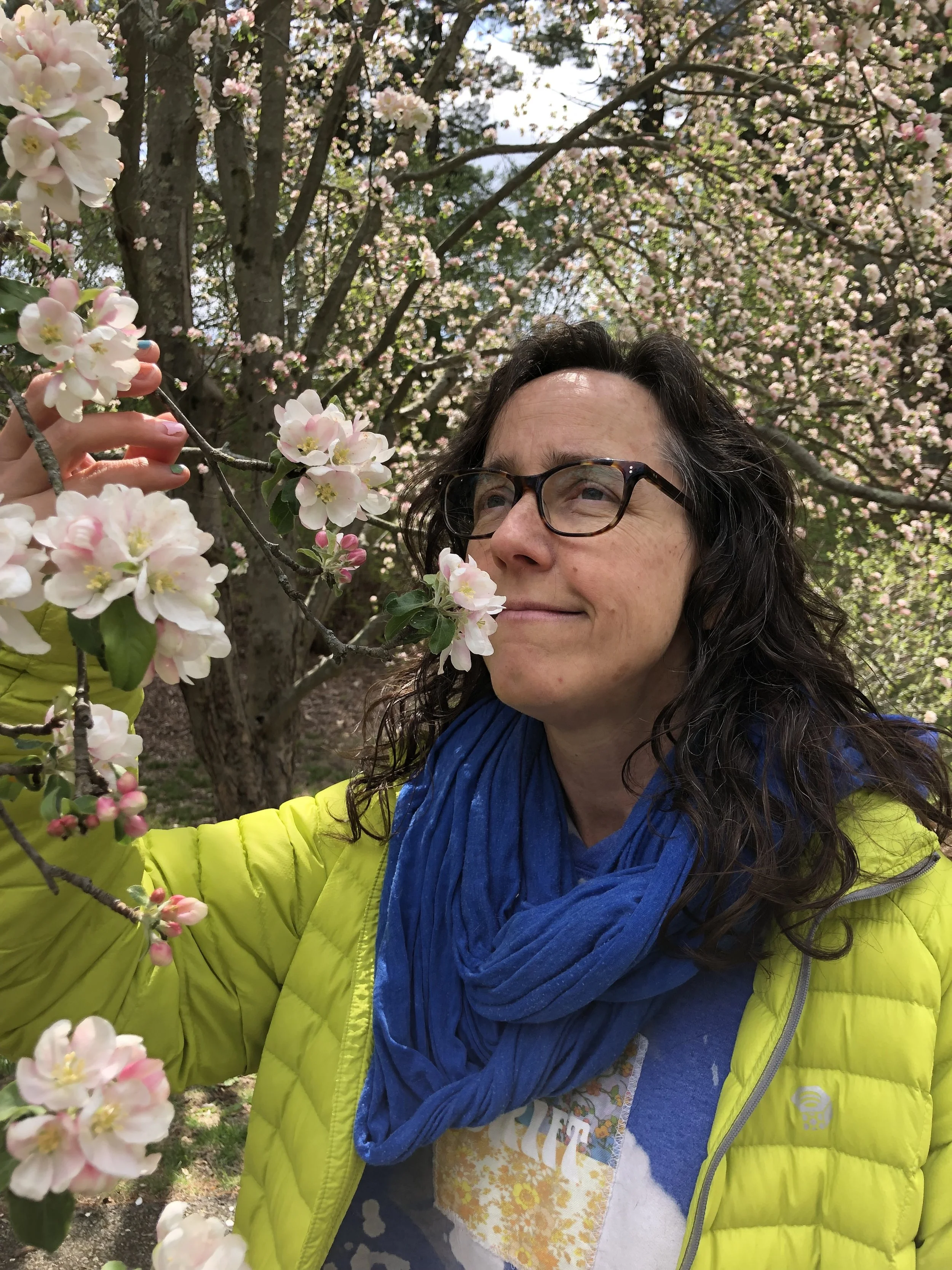 A woman with long brown hair and brown glasses holds a branch of crabapple blossoms close to her nose to take in the scent. She's wearing a yellow-green puffy coat and a bright blue circle scarf.The background is filled with more crabapple blossoms.