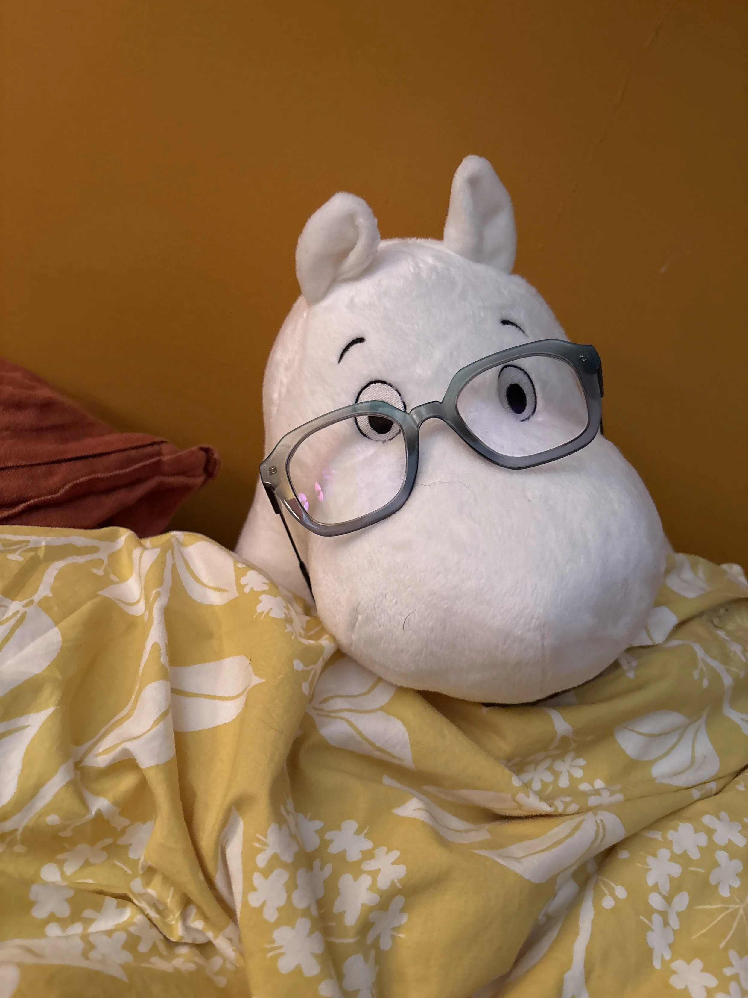 A white stuffed animal (Moomintroll) wears blue-grey glasses while tucked under a yellow, flowery blanket.