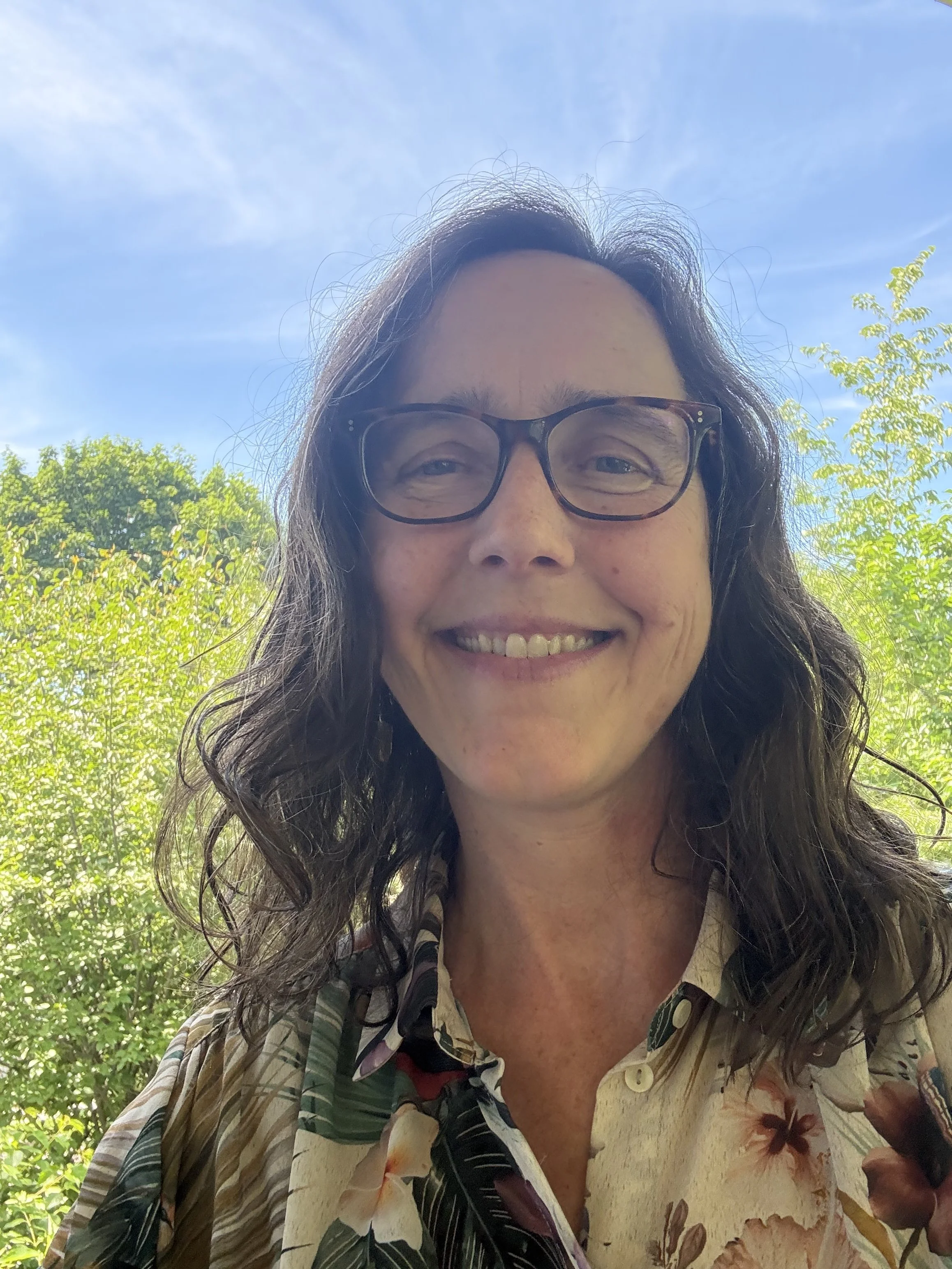 A woman with brown hair wearing glasses and a floral shirt, smiling outdoors with green trees and a blue sky in the background.