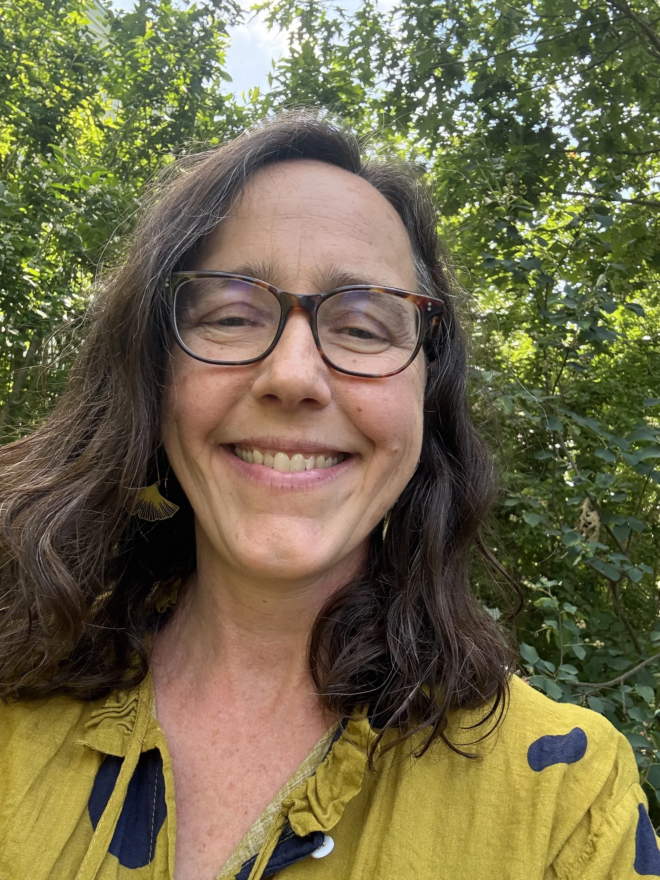 A smiling woman wit brown hair and glasses stands outside wearing a yellow blouse with large navy blue circles and ginkgo earrings