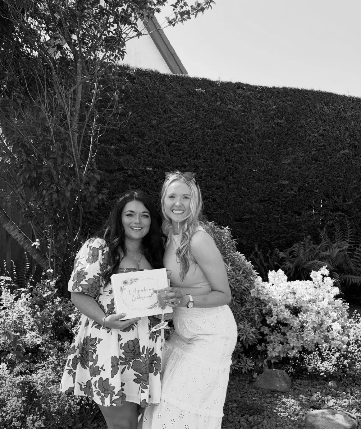 Two women standing outdoors in front of bushes and trees, smiling, holding a gift box and a glass of wine.