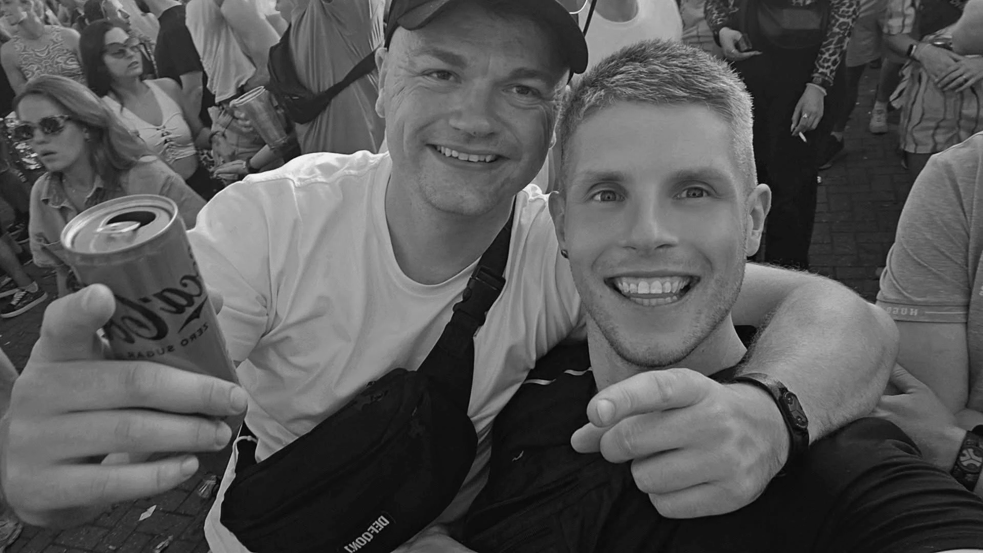 Two men smiling and taking a selfie at a crowded outdoor event, with other people in the background.
