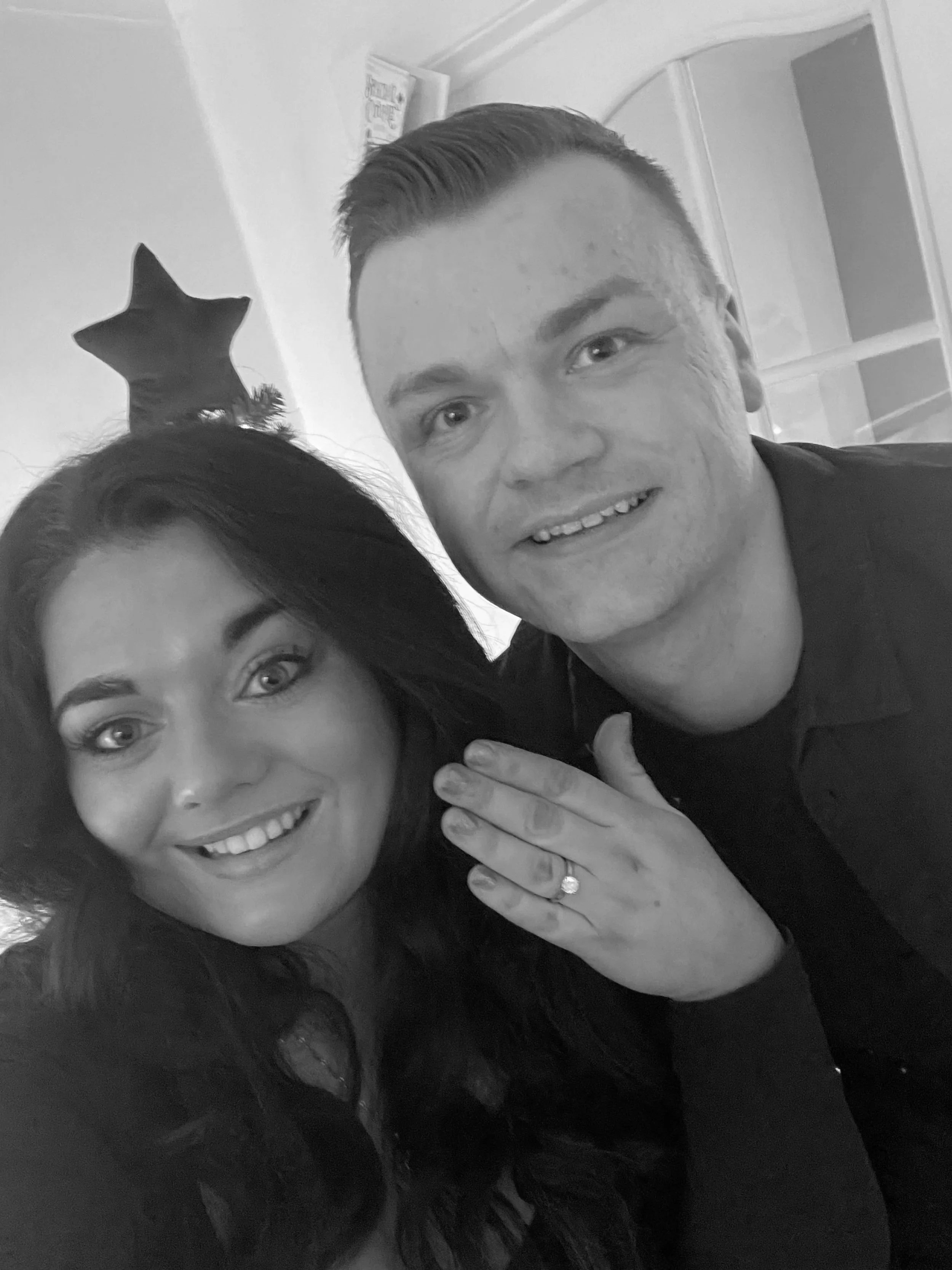 A black and white photo of a smiling woman and a smiling man close together, with the woman's left hand on the man's shoulder displaying a ring, and the woman is wearing a headband with a star on top.