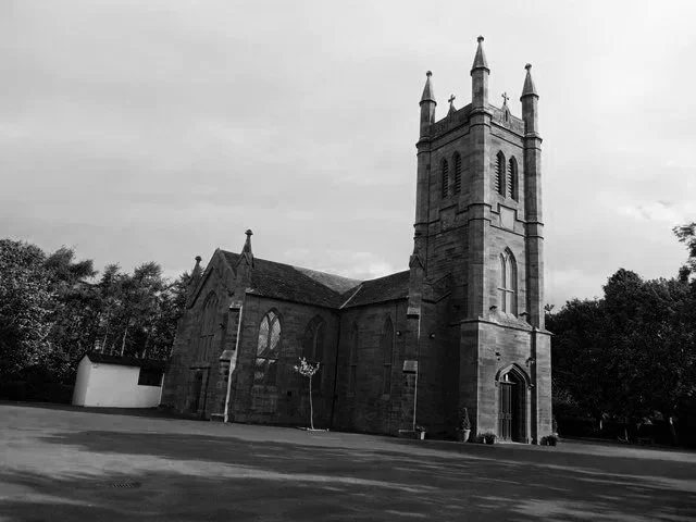 A black and white photo of a historic church with a tall steeple, surrounded by trees and an open lawn.