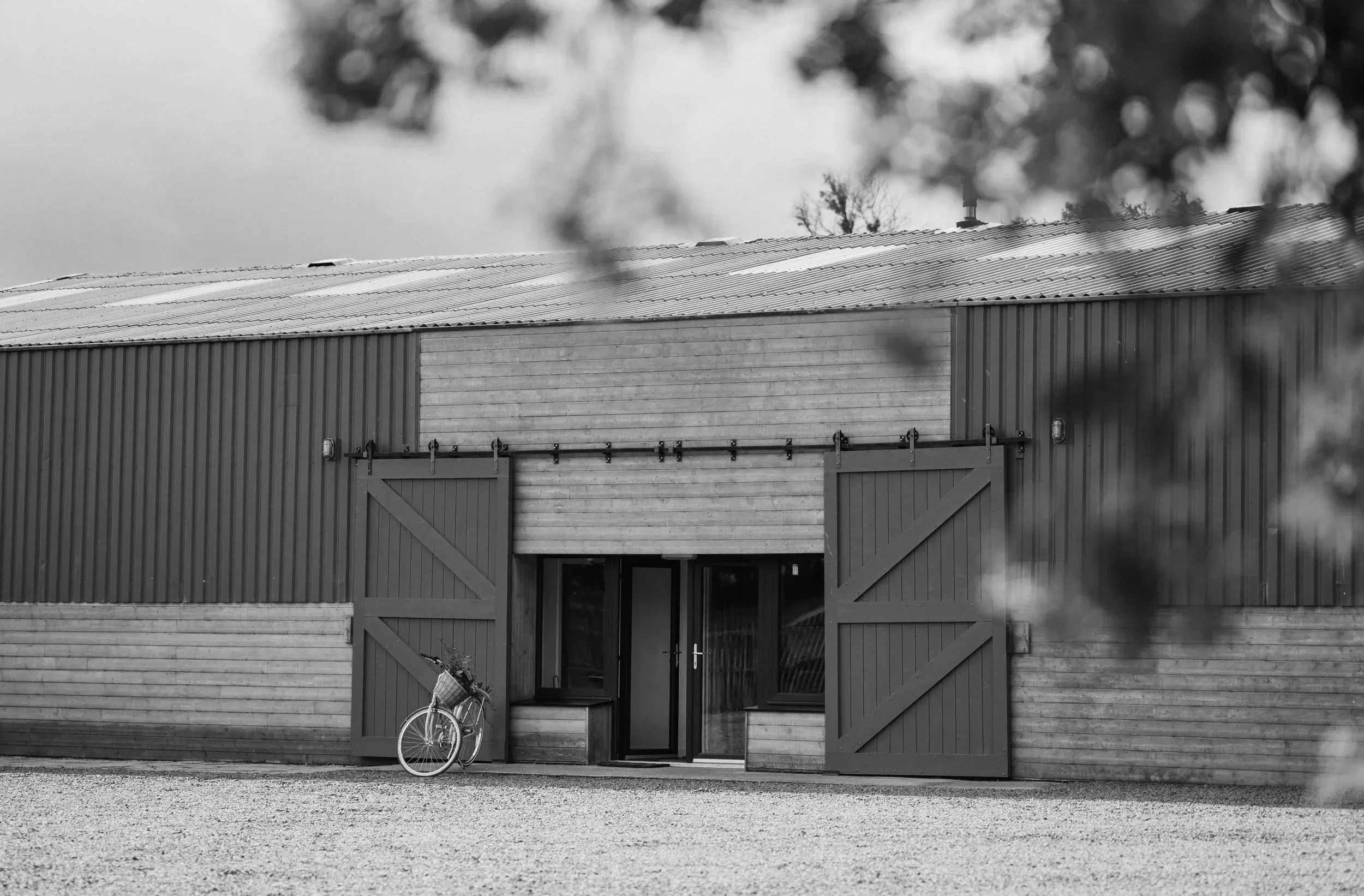 A black and white photo of a barn with large sliding doors, a bicycle leaning against the left side, and a gravel ground in front. The barn has a metal roof and wooden siding, with some trees and cloudy sky in the background.
