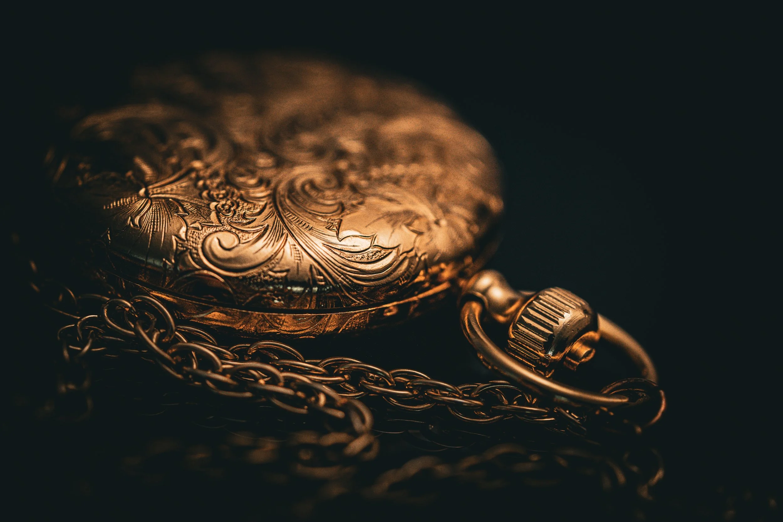 Close-up of an ornate, vintage gold pocket watch with intricate engravings, lying on a dark surface with a chain attached.