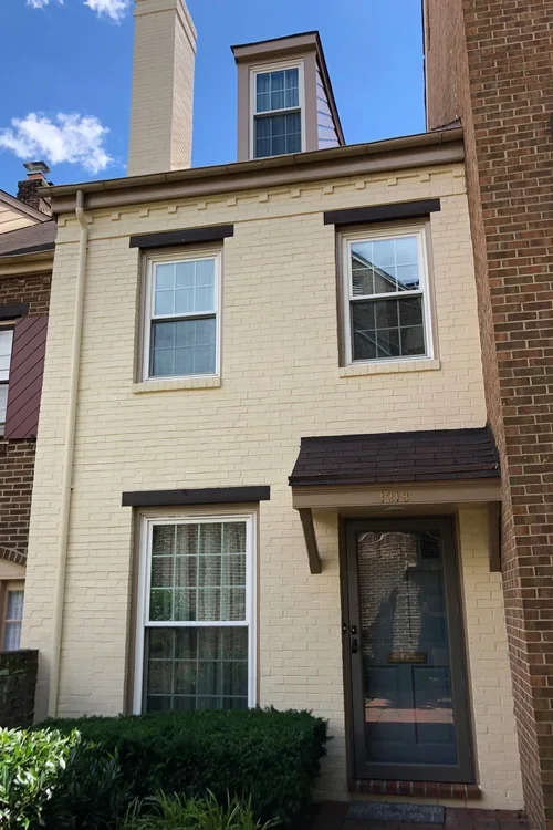 A three-story townhouse with a cream brick facade, black window frames, a black front door with a glass panel, a small awning above the door, and a second-floor dormer window.