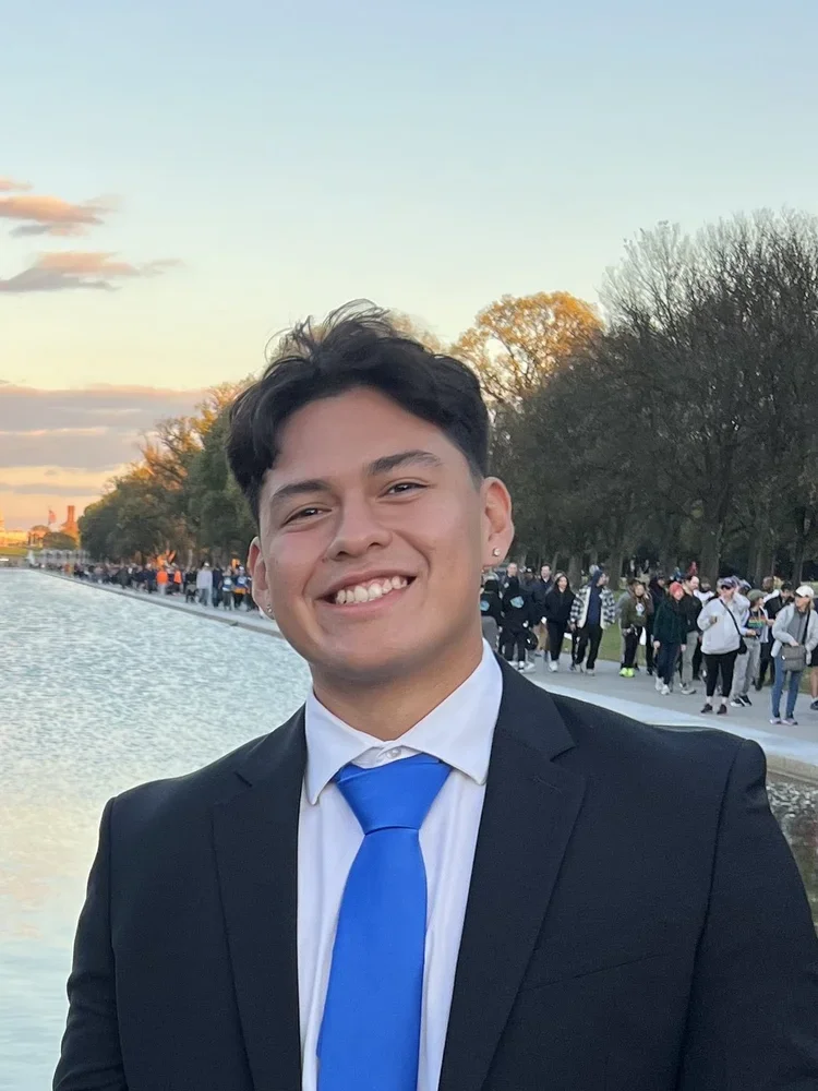 Young man in a suit with a blue tie smiling outdoors during the daytime, with a water body and a crowd of people walking along a park path in the background.