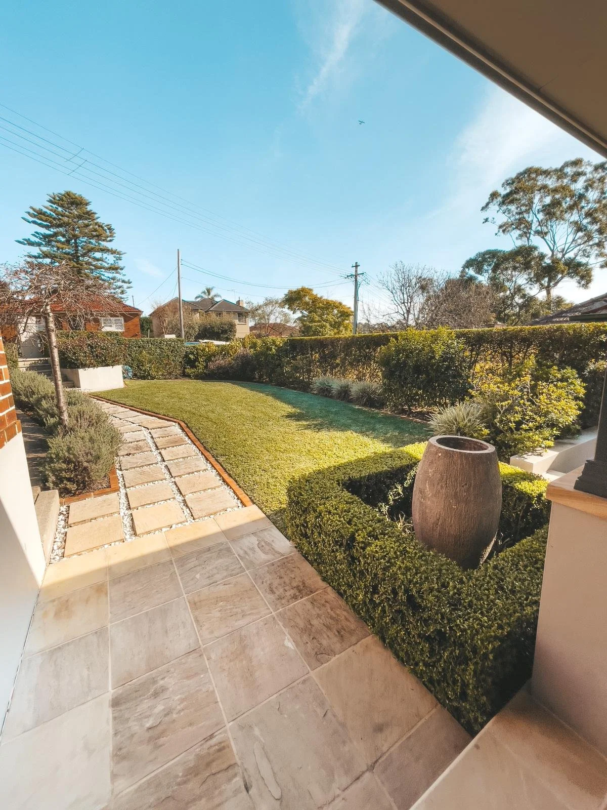 View of a backyard with a stone pathway, manicured lawn, shrubbery, trees, and blue sky.