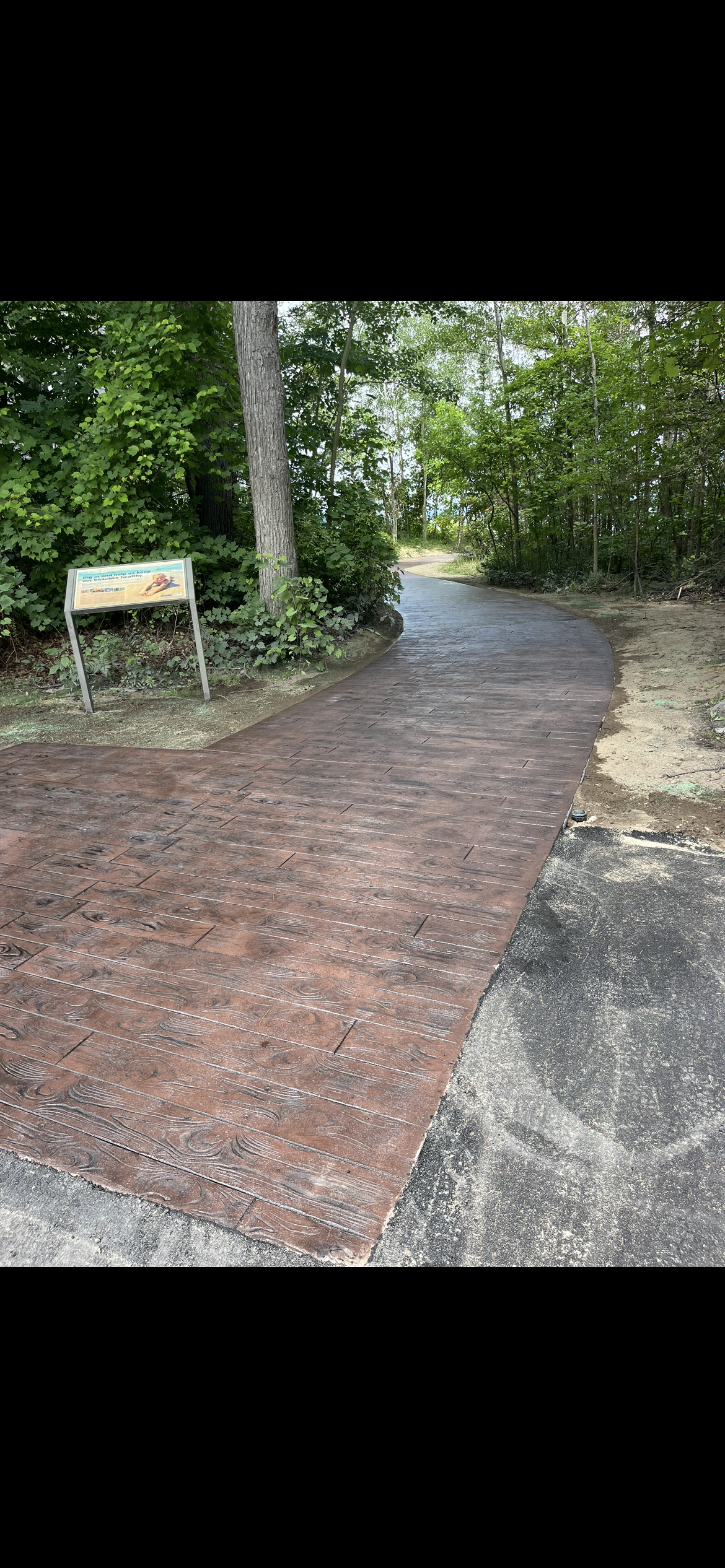 A paved walkway winding through a wooded area, with trees and greenery on both sides, and a signboard on the left side of the path.