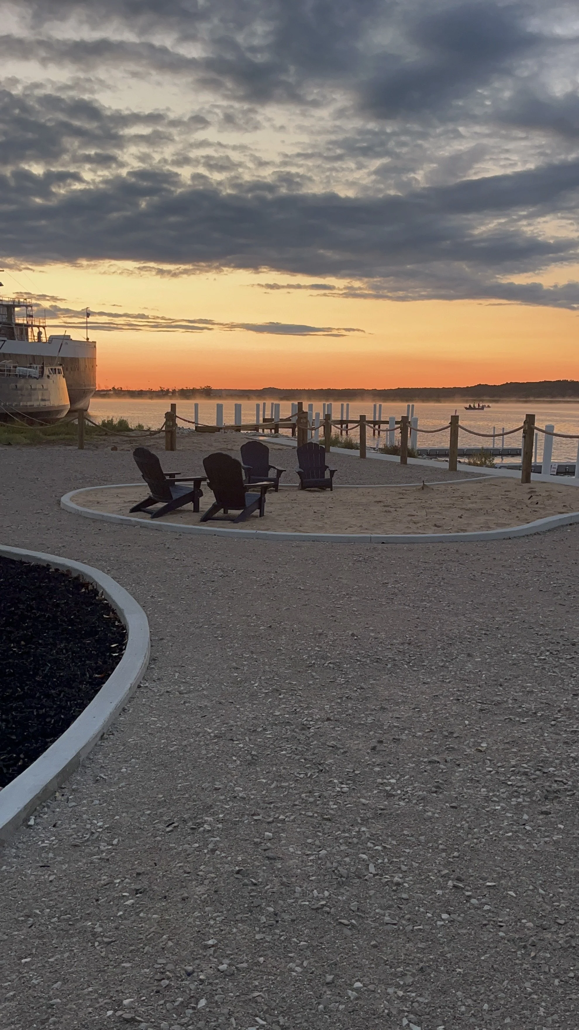 Sunset over a river with boats, a sandy area with four black Adirondack chairs, and a wooden fence along the water.