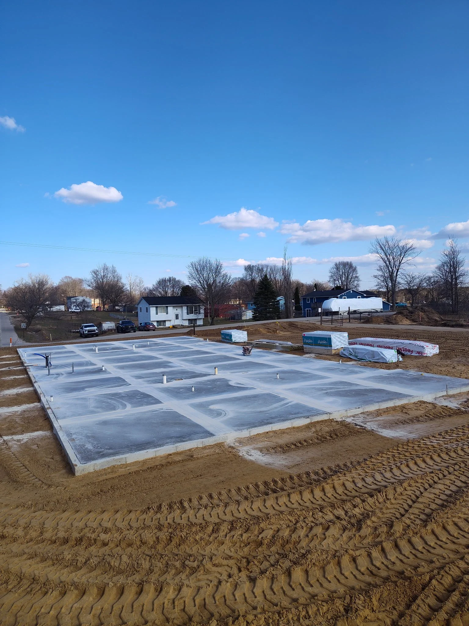 Construction site with concrete foundation and pipes, surrounded by dirt and tire tracks, under a blue sky with scattered clouds.