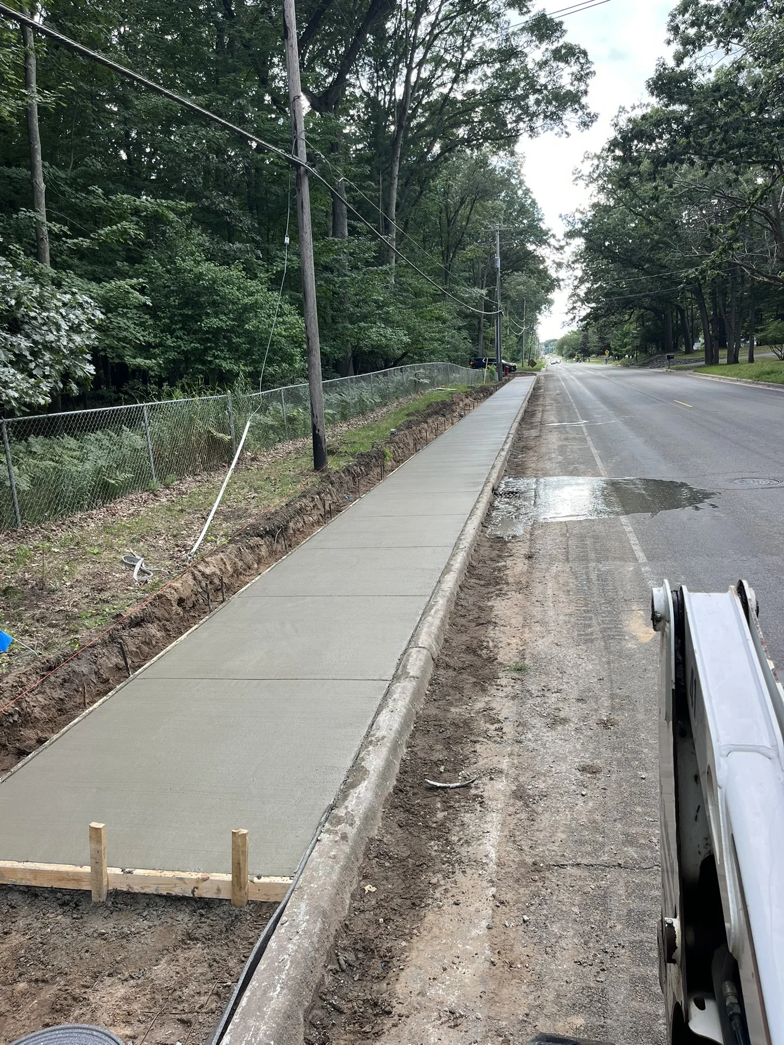 Newly poured concrete sidewalk under construction along a tree-lined street, with a construction vehicle and utility poles visible.