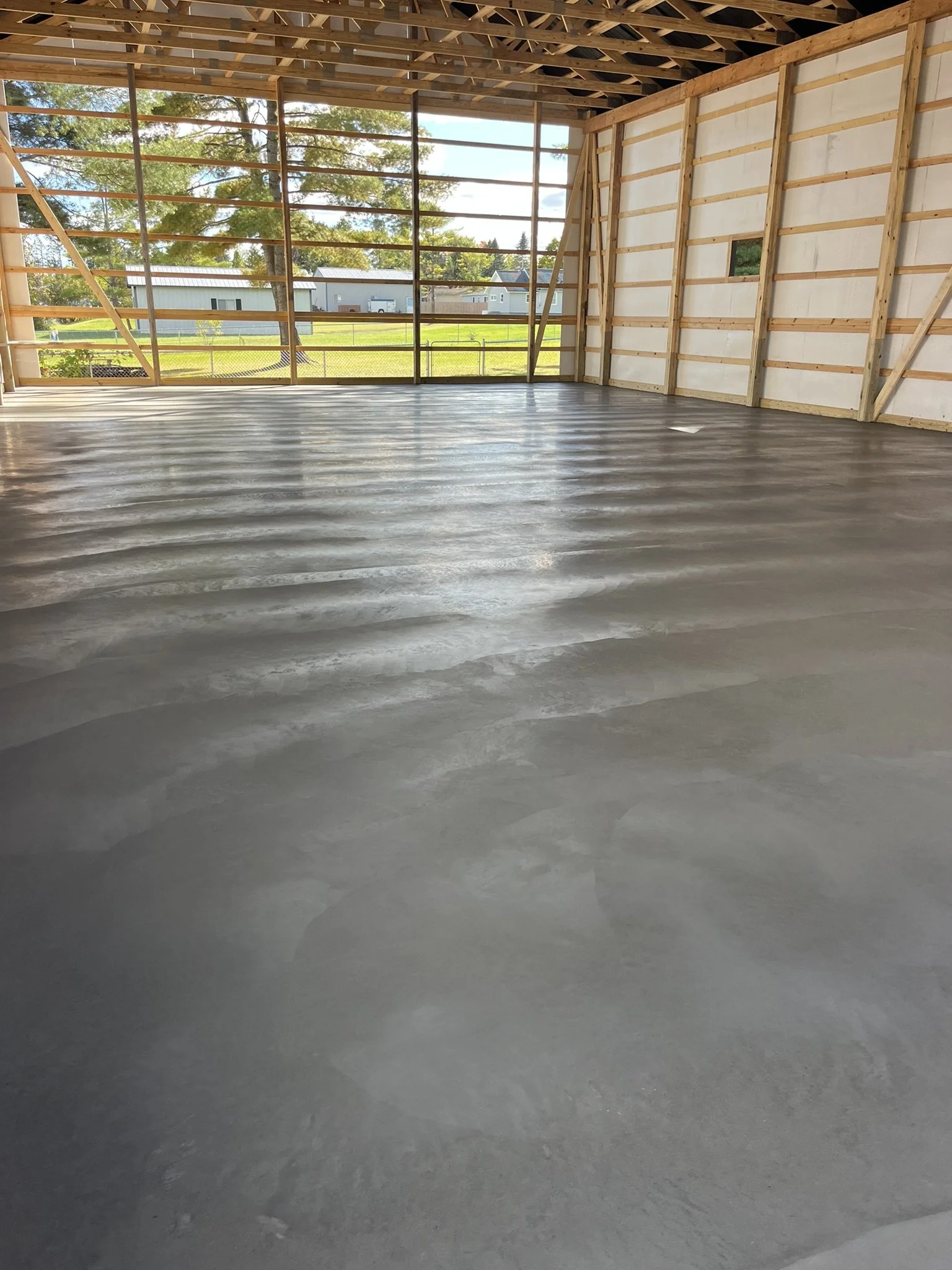 Inside a newly built wooden structure with open walls, showing a freshly poured concrete floor and large open window space revealing trees and a field outside.