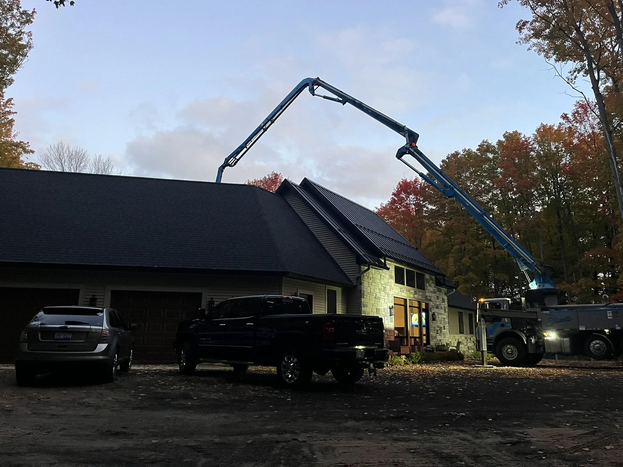 A house with solar panels on the roof being installed by a crane with a bucket arm, parked cars in the driveway, trees with fall foliage, and a cloudy sky at dusk.