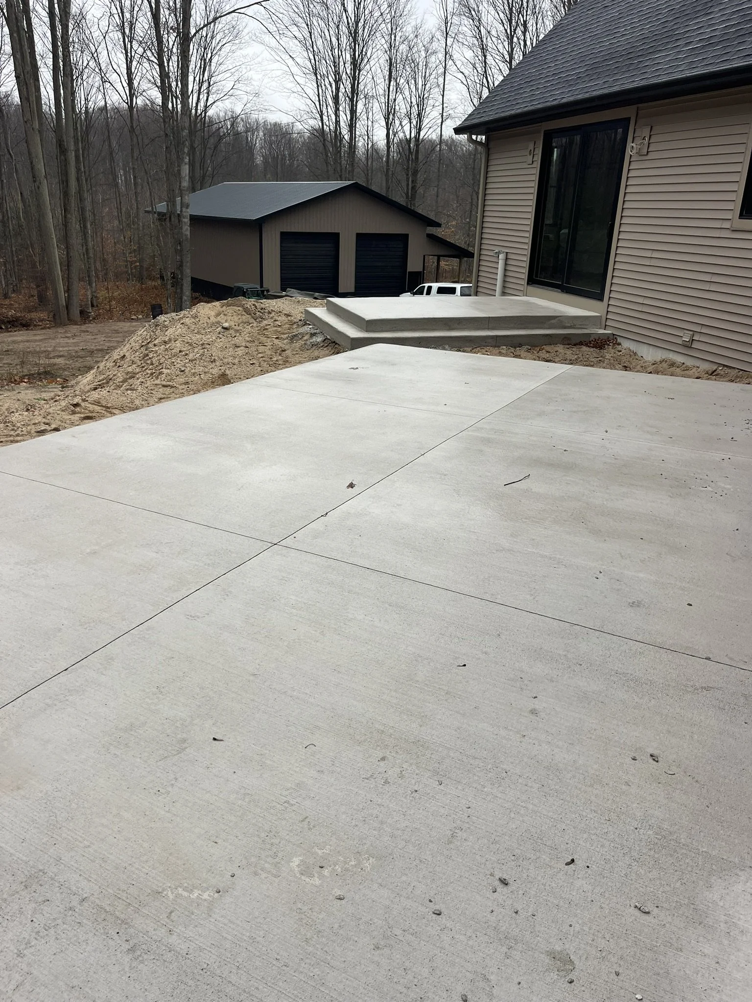 Newly poured concrete patio and steps outside a house, with a wooded backyard and a detached garage in the background.