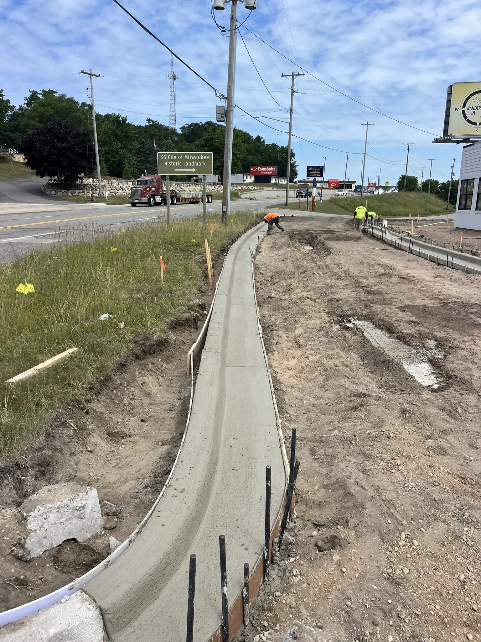 Construction workers concrete the sidewalk along a street with power lines, a grassy area, and signs for Milwaukee and a landmark in the background.