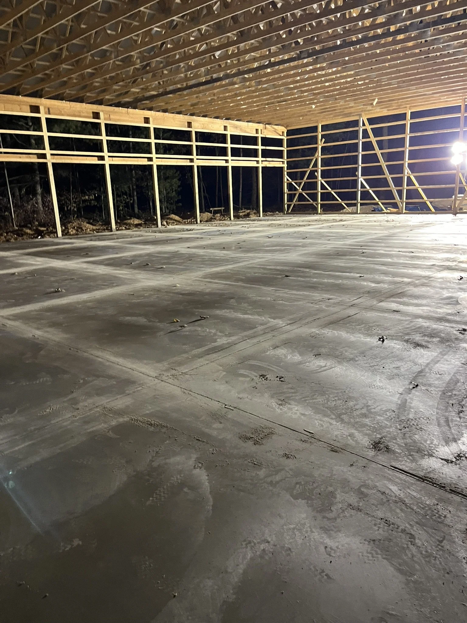 Construction site with a partially built wooden structure and a concrete floor, illuminated by a bright light at night.