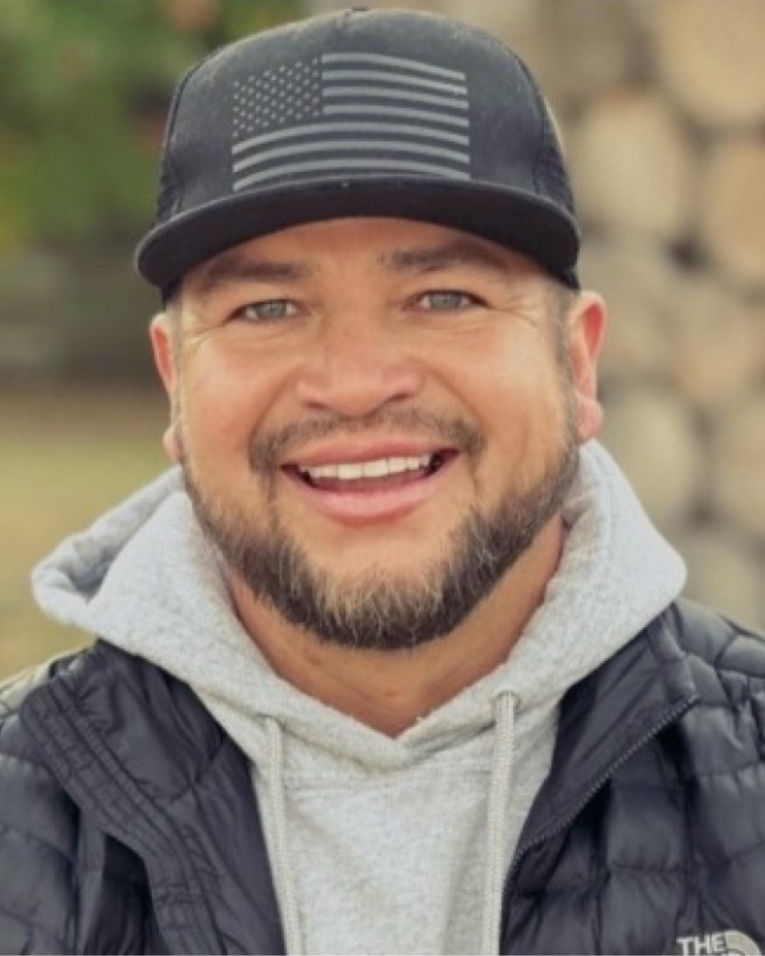 A smiling man with a beard and mustache wearing a black baseball cap with an American flag design, a gray hoodie, and a black puffer jacket outdoors.