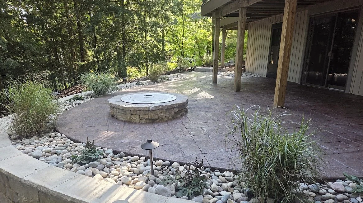 Backyard patio with a central fire pit, surrounded by decorative stones and lush greenery, connected to a house with a covered patio area.