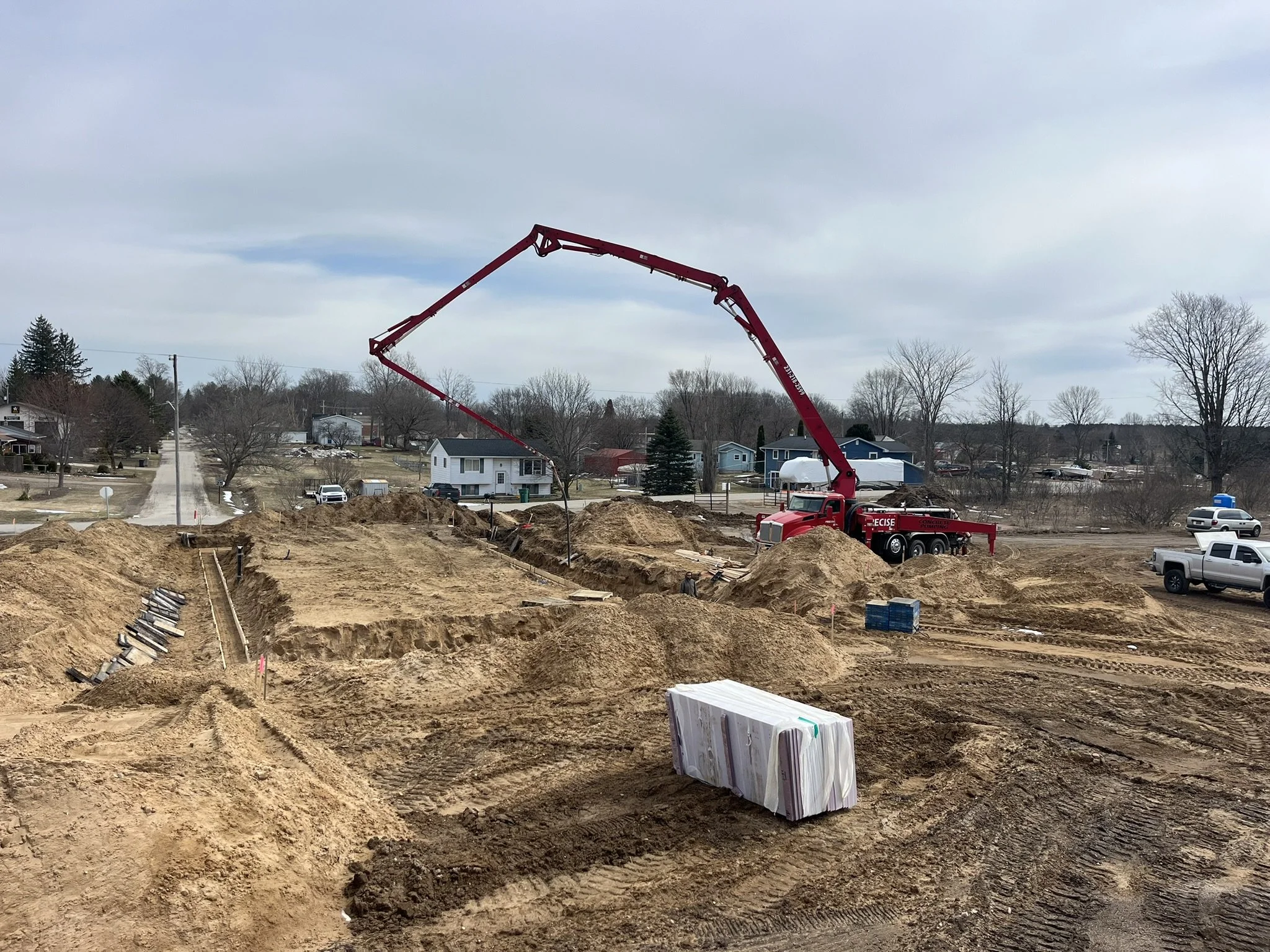 Construction site with excavated earth, a red concrete pump truck with extended arm, several piles of dirt, and construction materials and vehicles, with residential houses and leafless trees in the background under a cloudy sky.