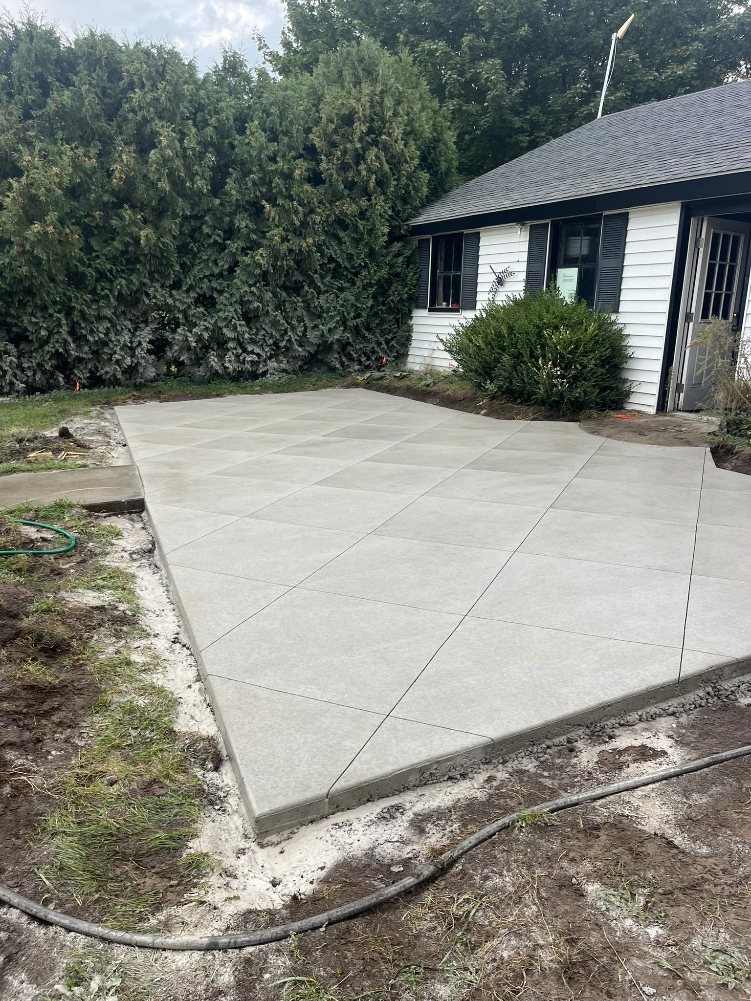 Newly poured concrete patio with large square tiles adjacent to a white house with black shutters, surrounded by green lawn and bushes.