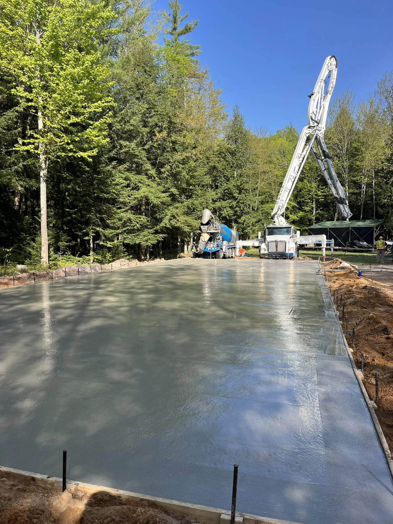 Concrete being poured on a construction site surrounded by trees, with a concrete pump truck and a cement mixer truck in the background.