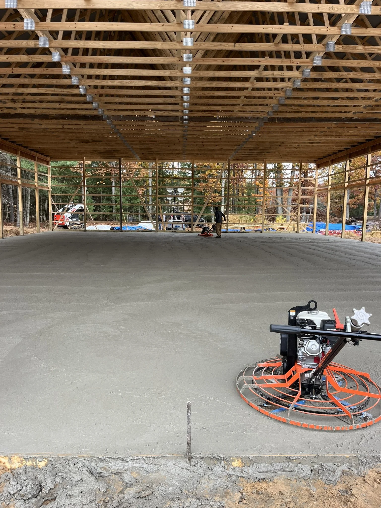 Construction site with a partially built wooden structure and a large concrete slab floor. A worker is operating a power trowel on the floor, smoothing the concrete. The background shows trees and construction equipment.