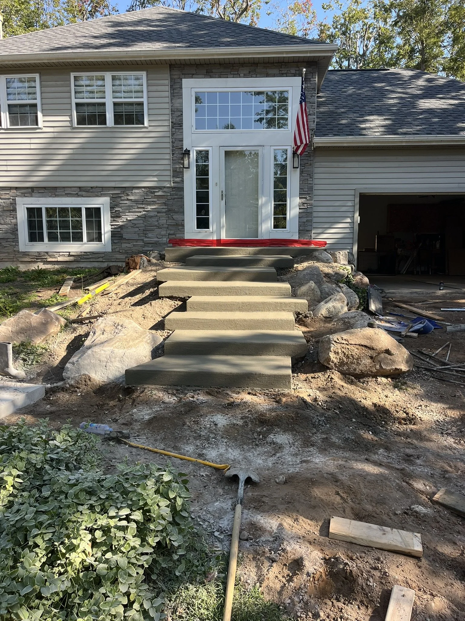 Front entrance of a house under construction with new concrete steps, large rocks on each side, an American flag on the right, and interior of the garage visible on the right.