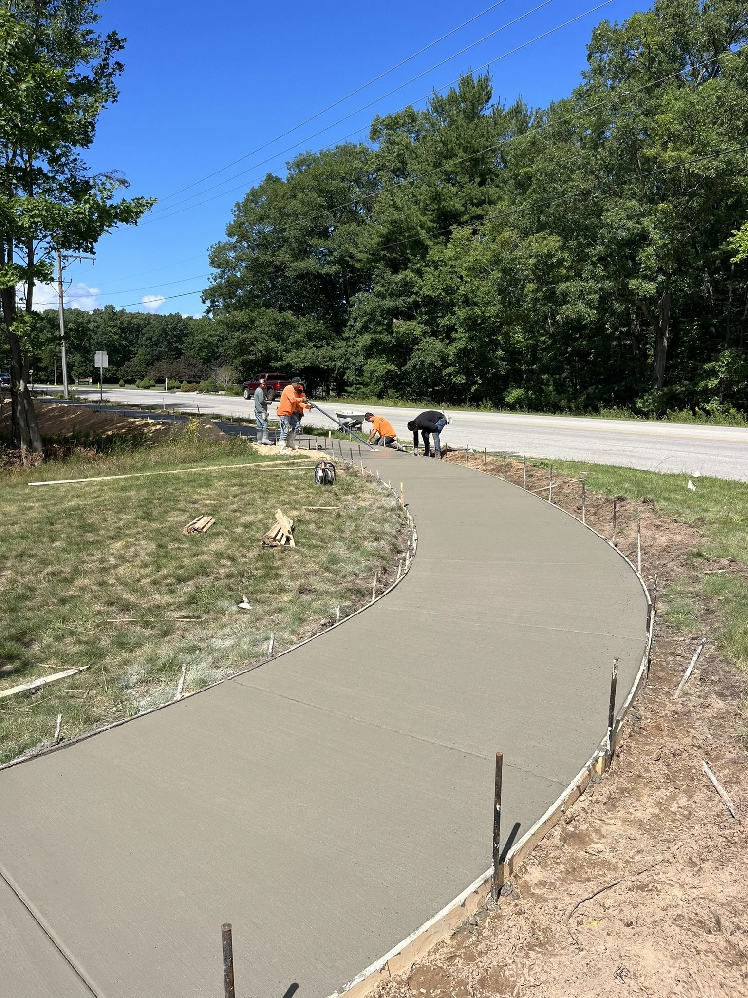 Workers pouring and smoothing concrete for a sidewalk along a grassy area next to a road, with trees and power lines in the background.