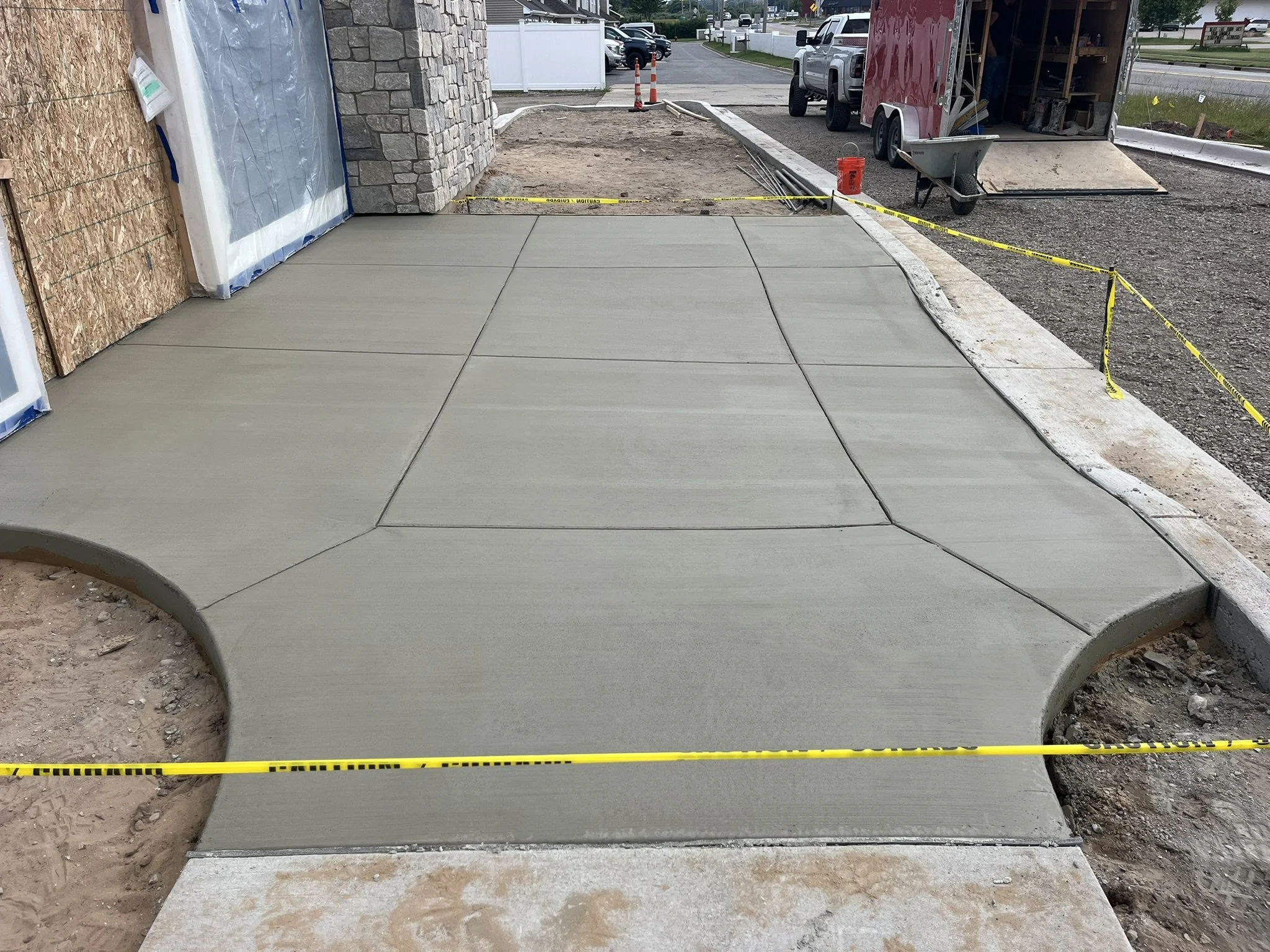 Freshly poured concrete sidewalk with sections outlined, red construction trailer with open door, construction tools and materials around, and caution tape preventing access.