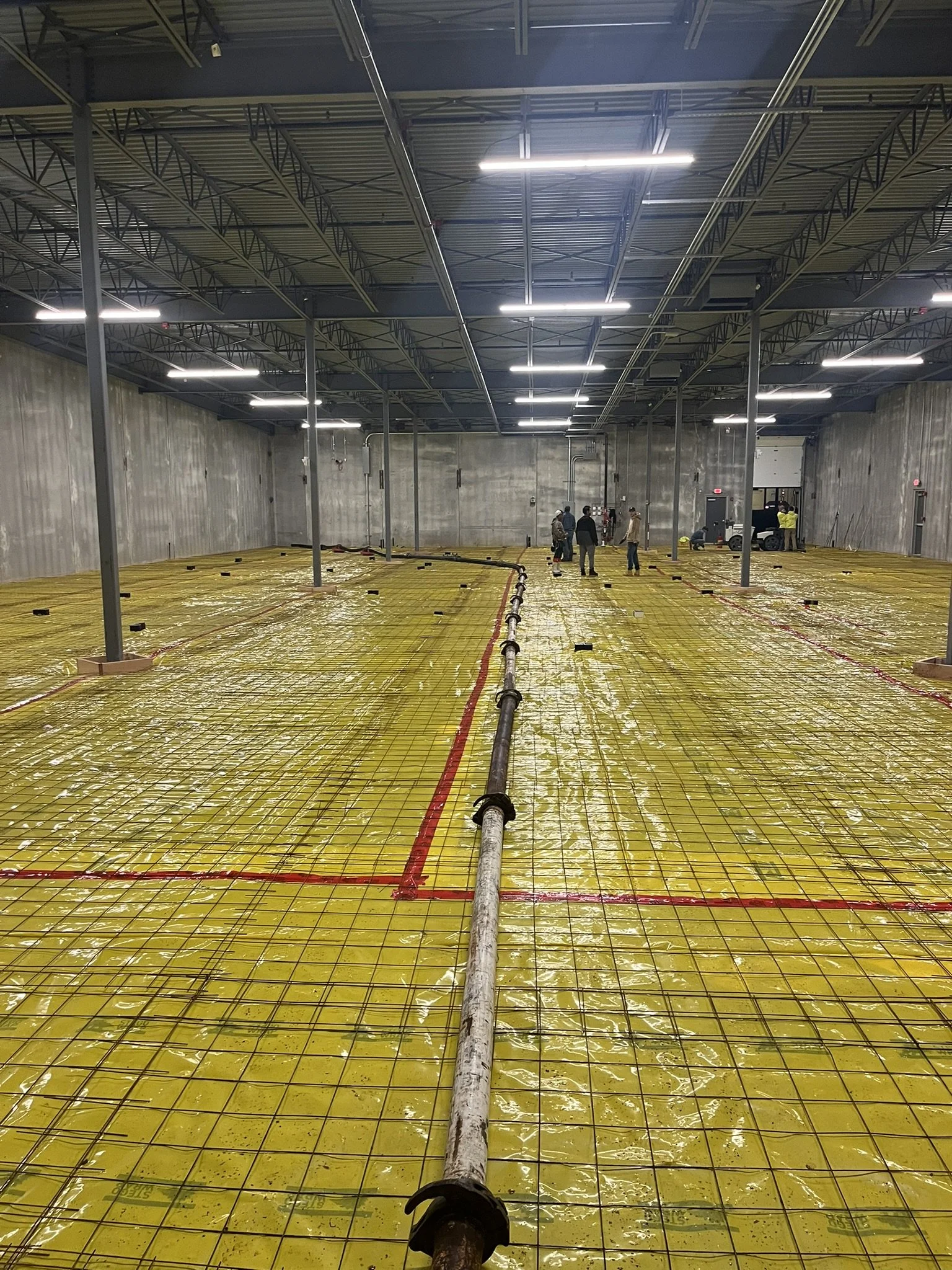 Construction workers in an underground structure with exposed concrete walls and ceiling, laying yellow insulation panels reinforced with metal rebar and a pipe running through the middle.